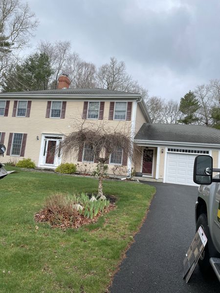 Two-story beige house with brown shutters and a weeping tree in front, on a green lawn with a black driveway.