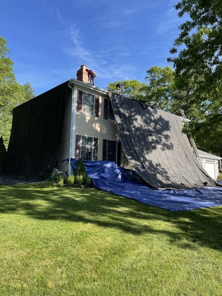 House covered in black tarp and blue tarp on lawn, likely for roof repair. Sunny day.