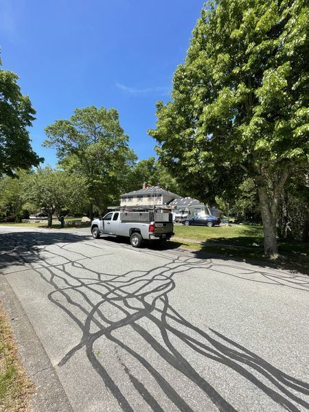 Silver truck parked on a road with visible black asphalt patches; green trees and house in the background under a blue sky.