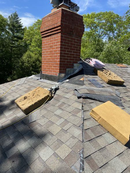 Rooftop with a brick chimney and damaged roofing shingles. Beige and brown hues, blue sky.