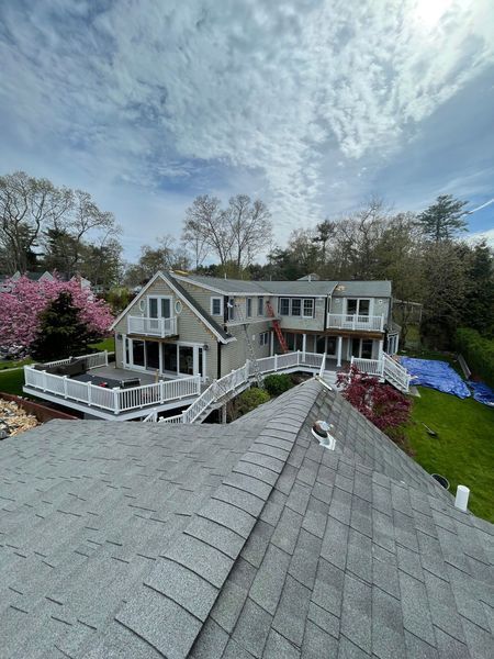 Gray house with white decks, under cloudy sky, surrounded by trees and a green lawn.