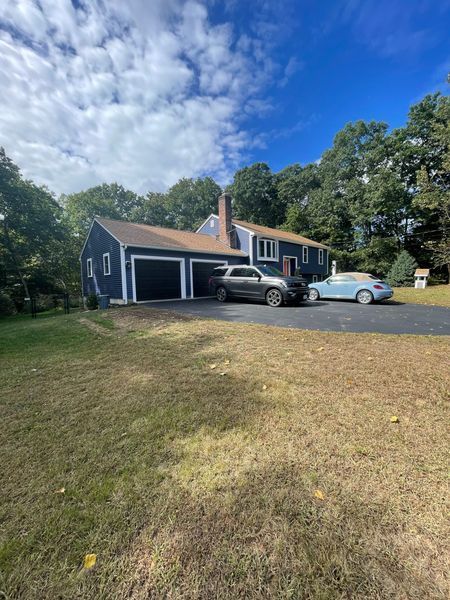Blue house with two-car garage, two cars in driveway, surrounded by trees and grass, under a blue sky.