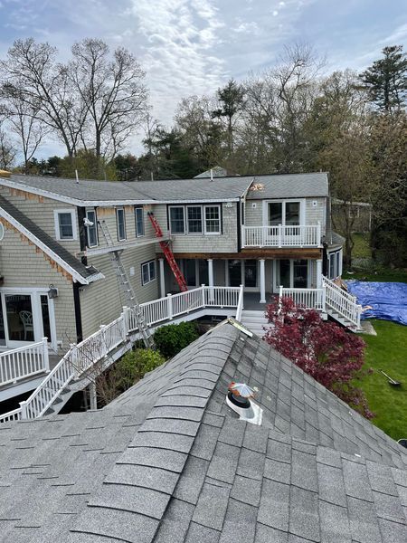 House with gray roof, undergoing repairs. Ladders, scaffolding, and blue tarp present. Overcast sky.