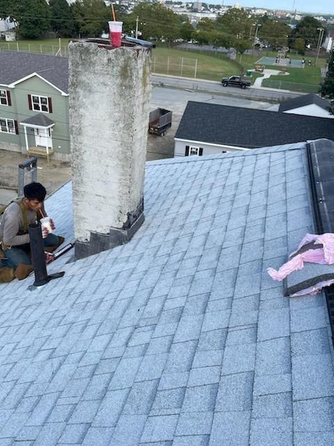 Roofer working on a gray shingle roof near a chimney. A child's clothing is on the roof, and a park is in the background.
