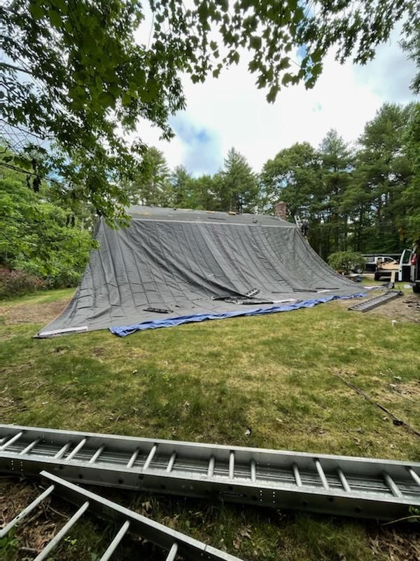 House roof covered with a large, dark tarp, grass, and a ladder in the foreground.
