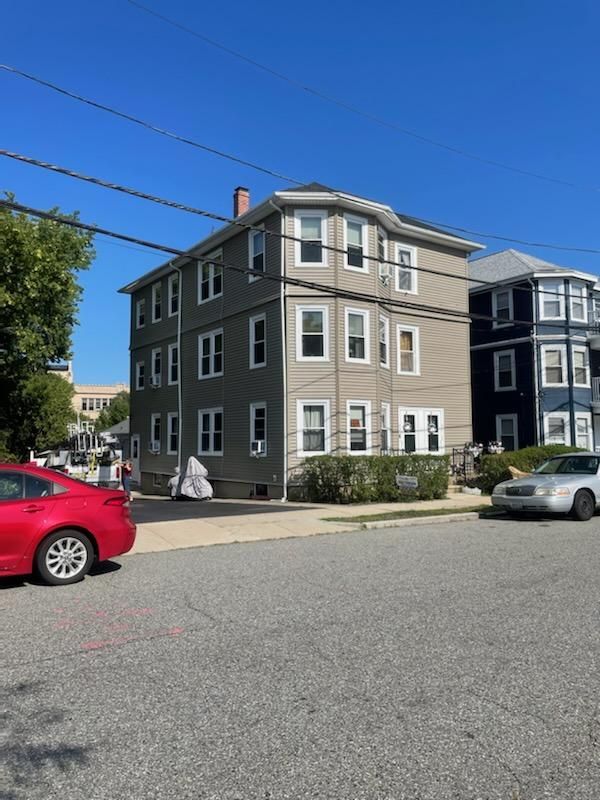 Three-story building with tan and gray siding on a city street; cars parked nearby on a sunny day.