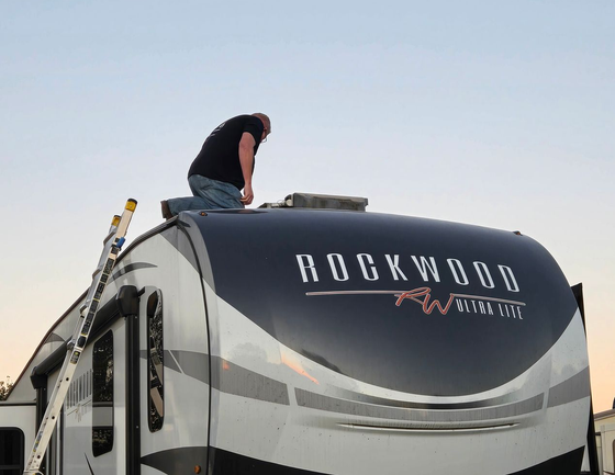 Person on RV roof near Rockwood logo, working with tools; ladder nearby, clear sky in background.