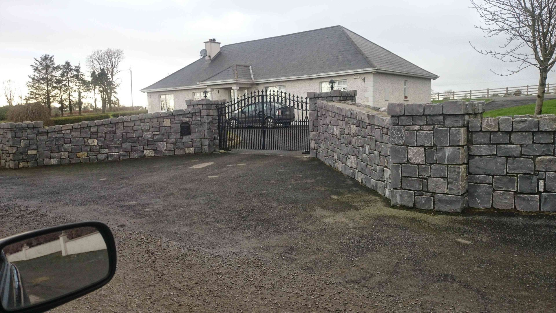 A stone wall with a metal gate leading to a light-colored house with a dark roof on a gravel driveway.