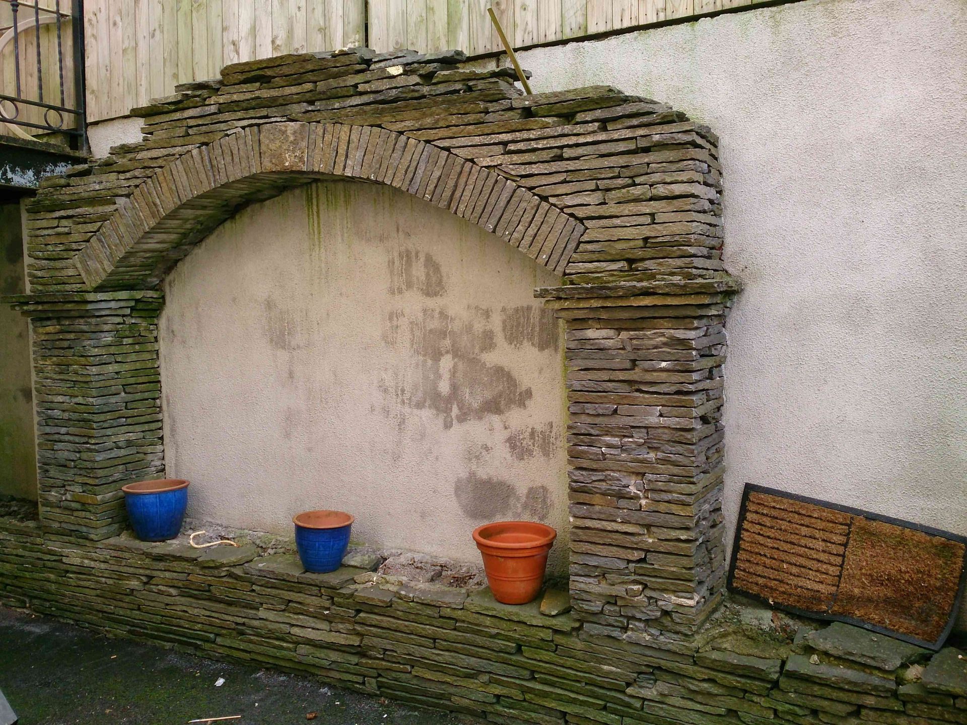A stone arch feature built against a stucco wall, with three colorful plant pots sitting on a ledge at its base.