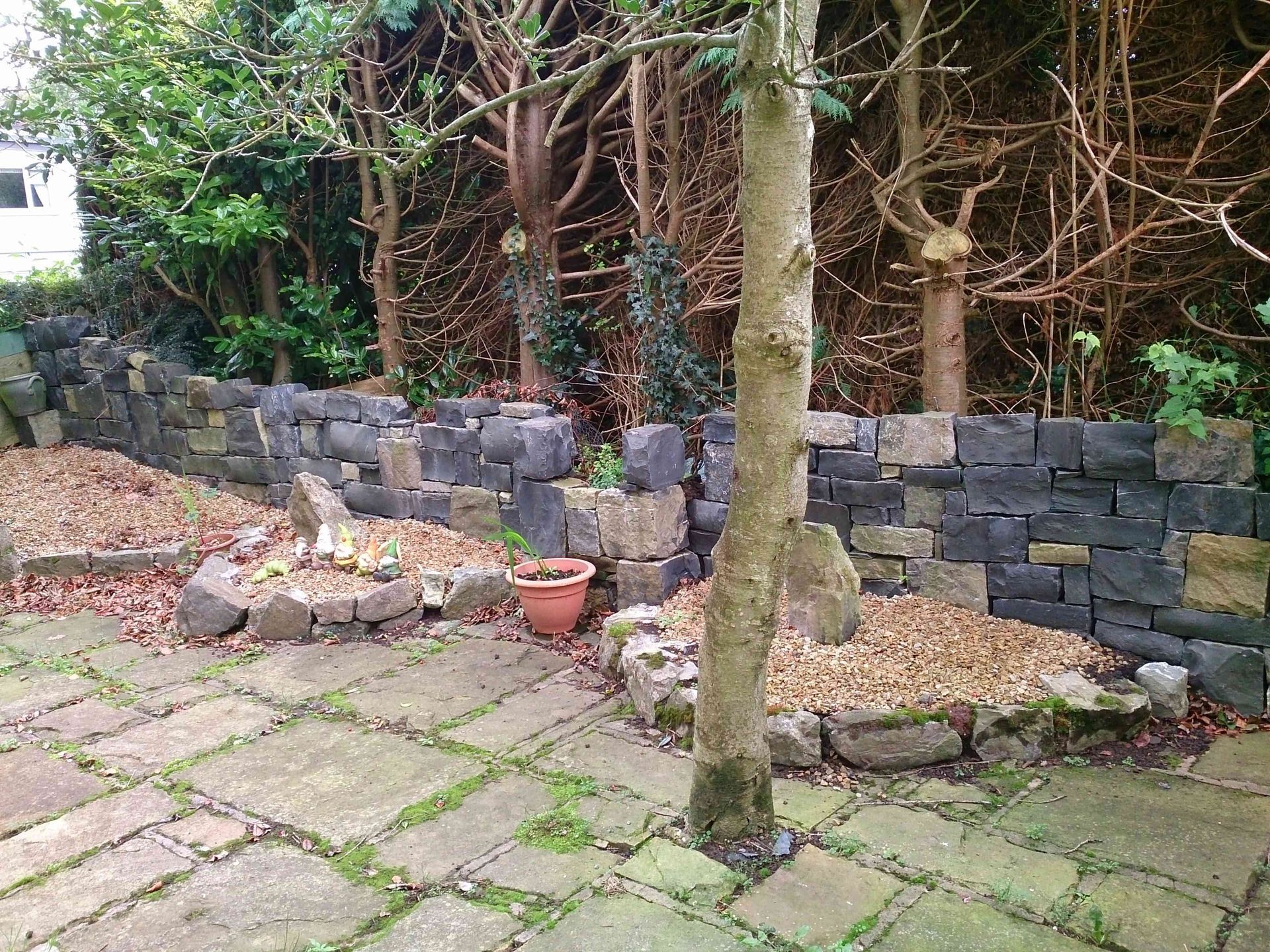 A stone retaining wall curves behind a patio, with trees and mulch-covered garden beds in front.