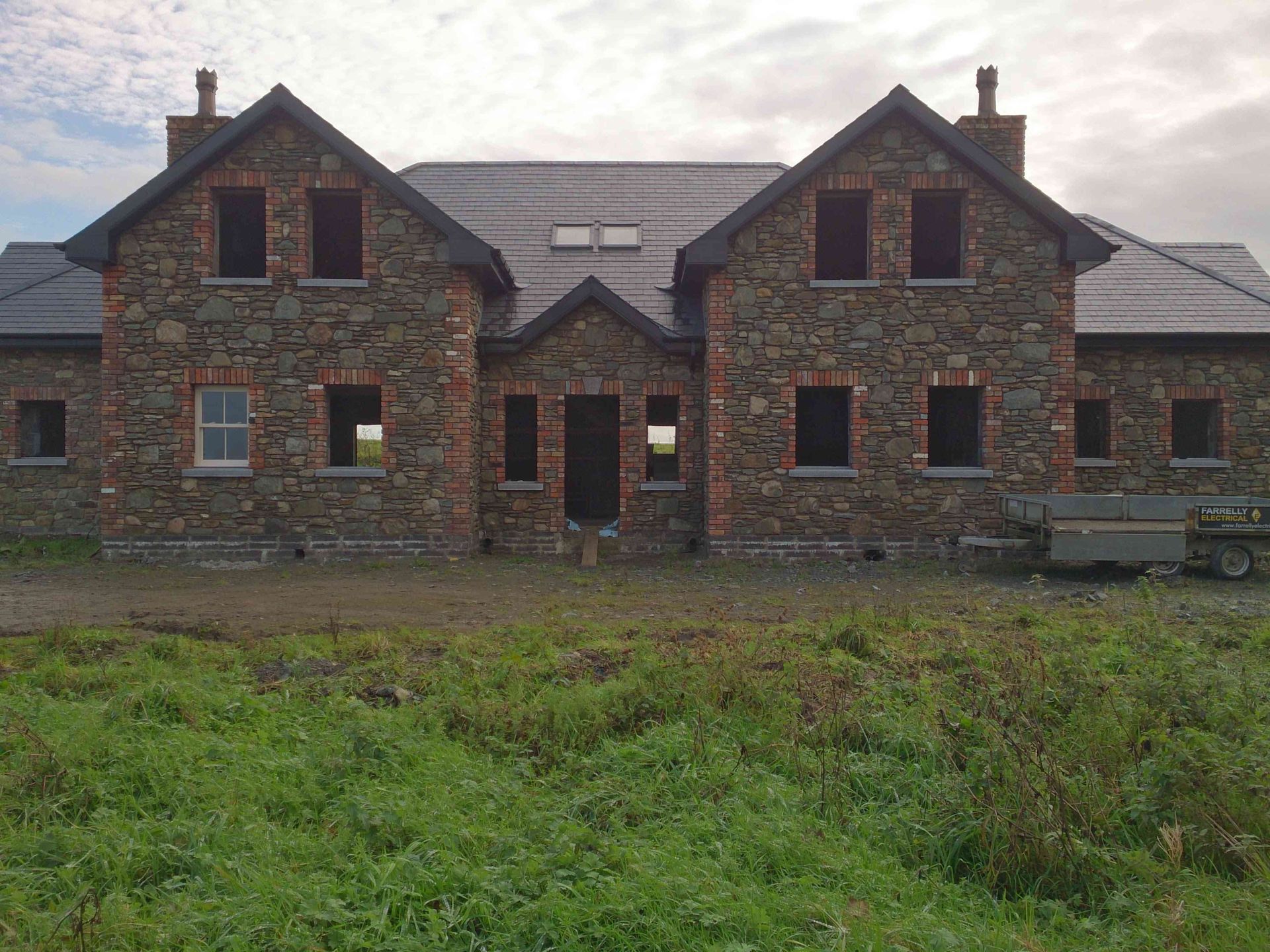 A two-story stone house under construction, with a symmetrical facade, two gables, and dark roof tiles in a grassy field.
