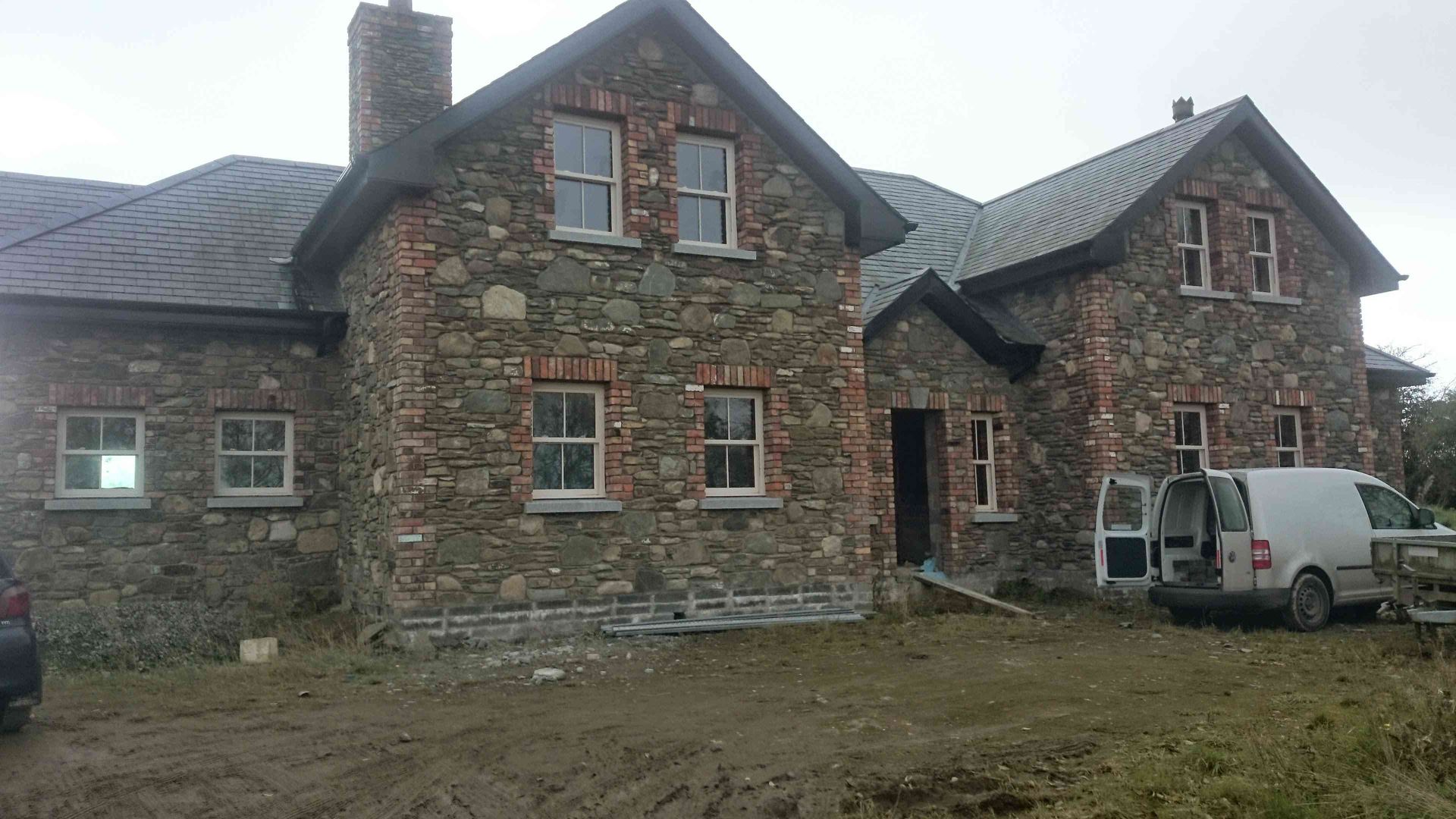 A two-story stone house under construction with a white van parked in front on a gravel lot.