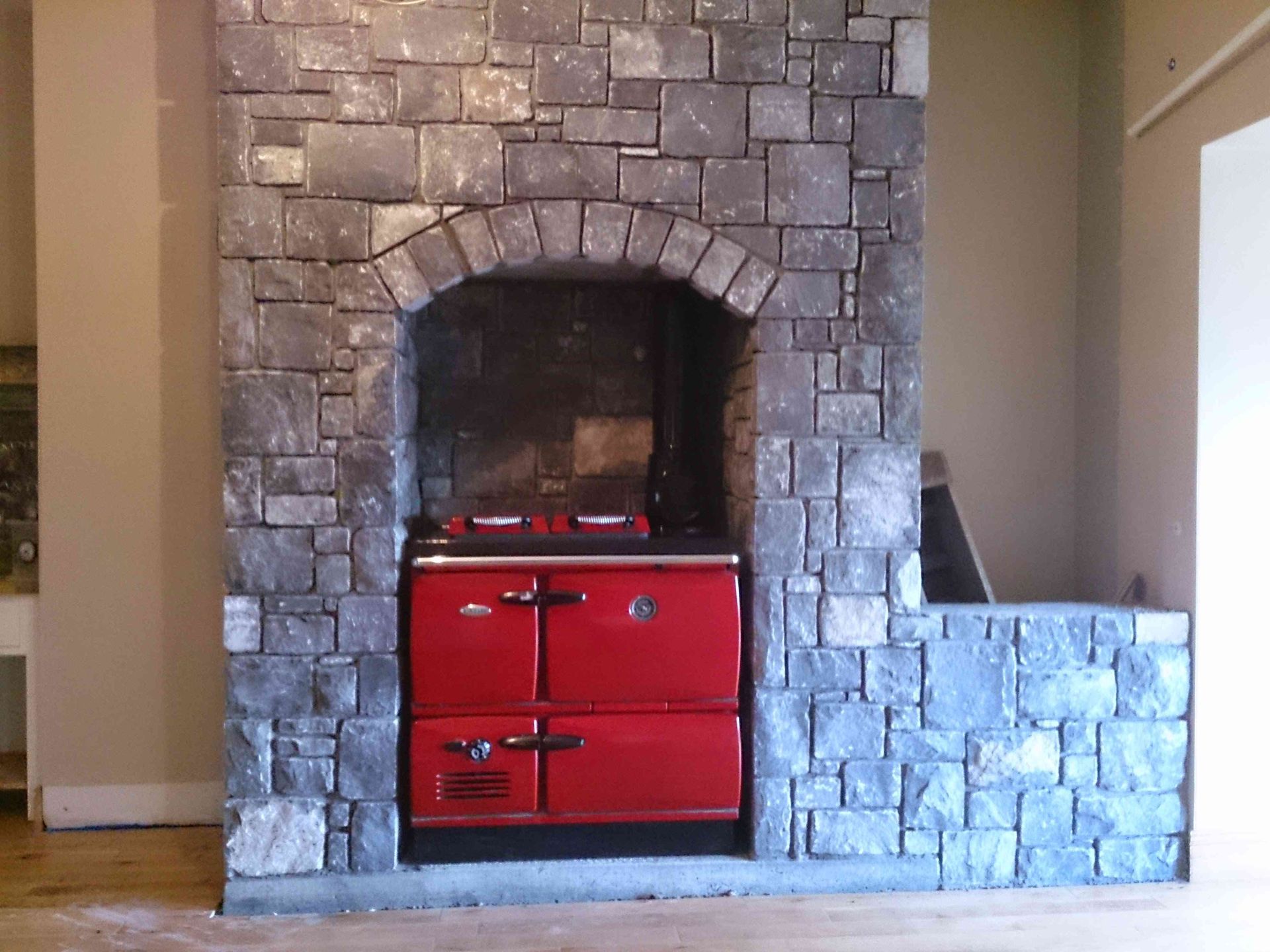 A bright red AGA cooker set inside a stone-clad kitchen fireplace alcove with a matching stone wall extending to the right.