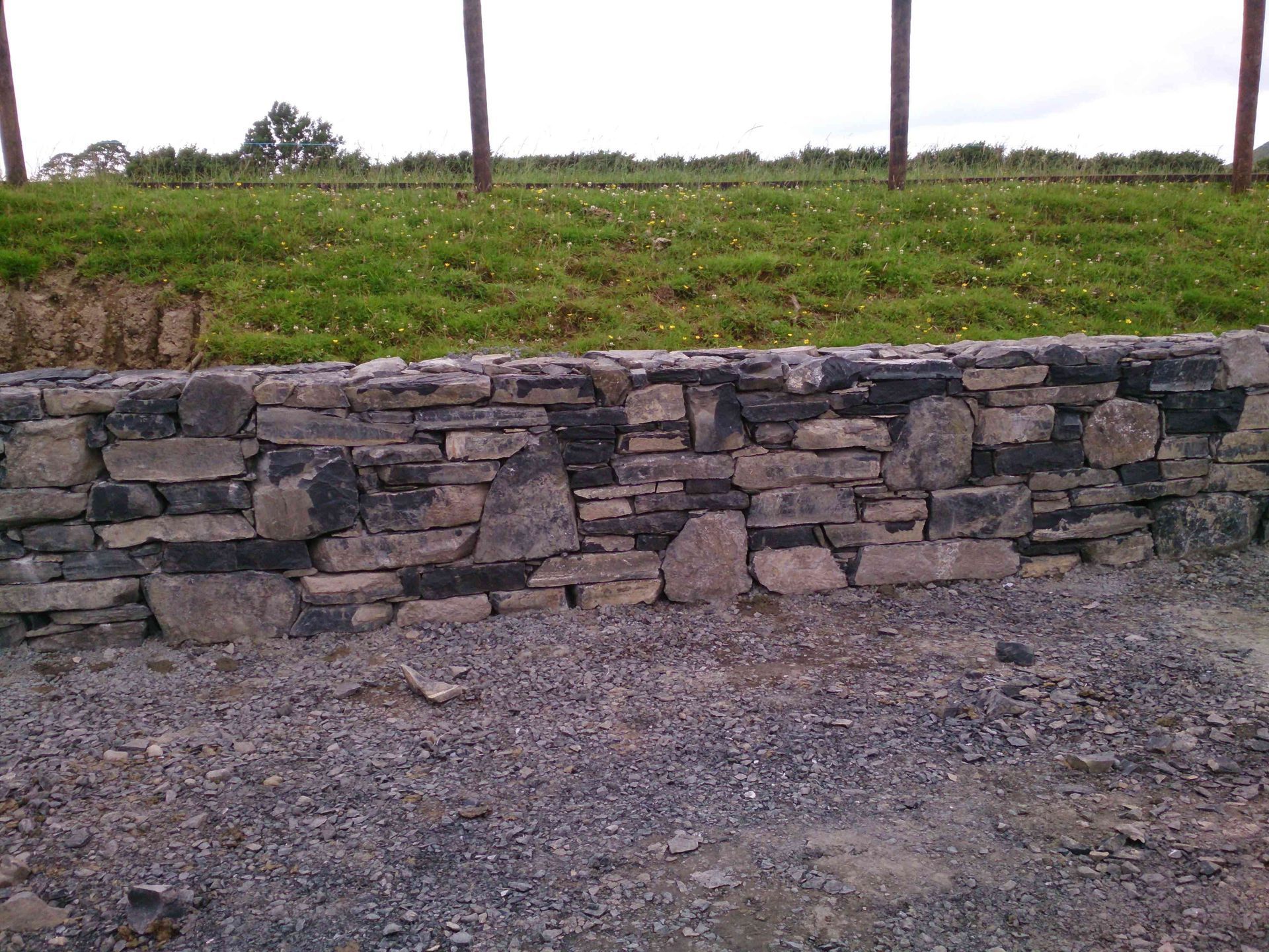 A dry-stone wall made of various gray and brown rocks, situated in front of a grassy hill with wooden fence posts.