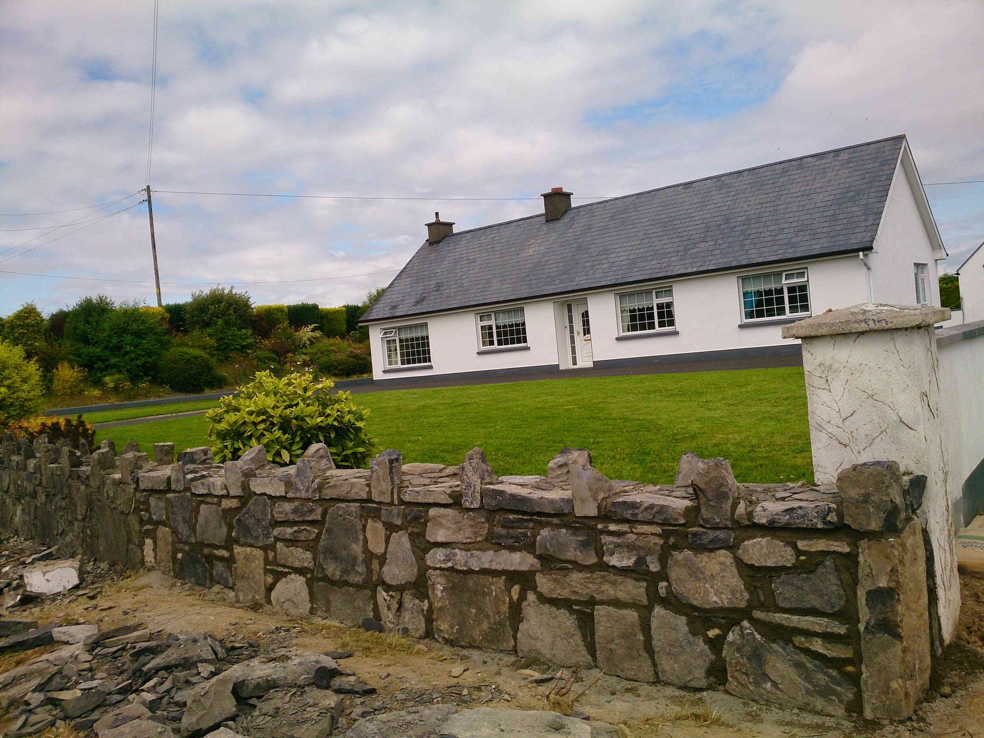 A white cottage with a dark grey roof behind a low, rustic stone wall and a green lawn under a cloudy sky.