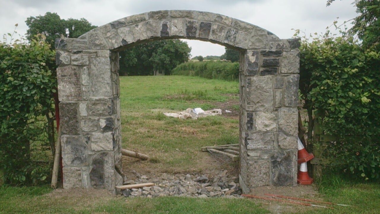 A stone archway stands between two green hedges, leading into a grassy field under a cloudy sky.