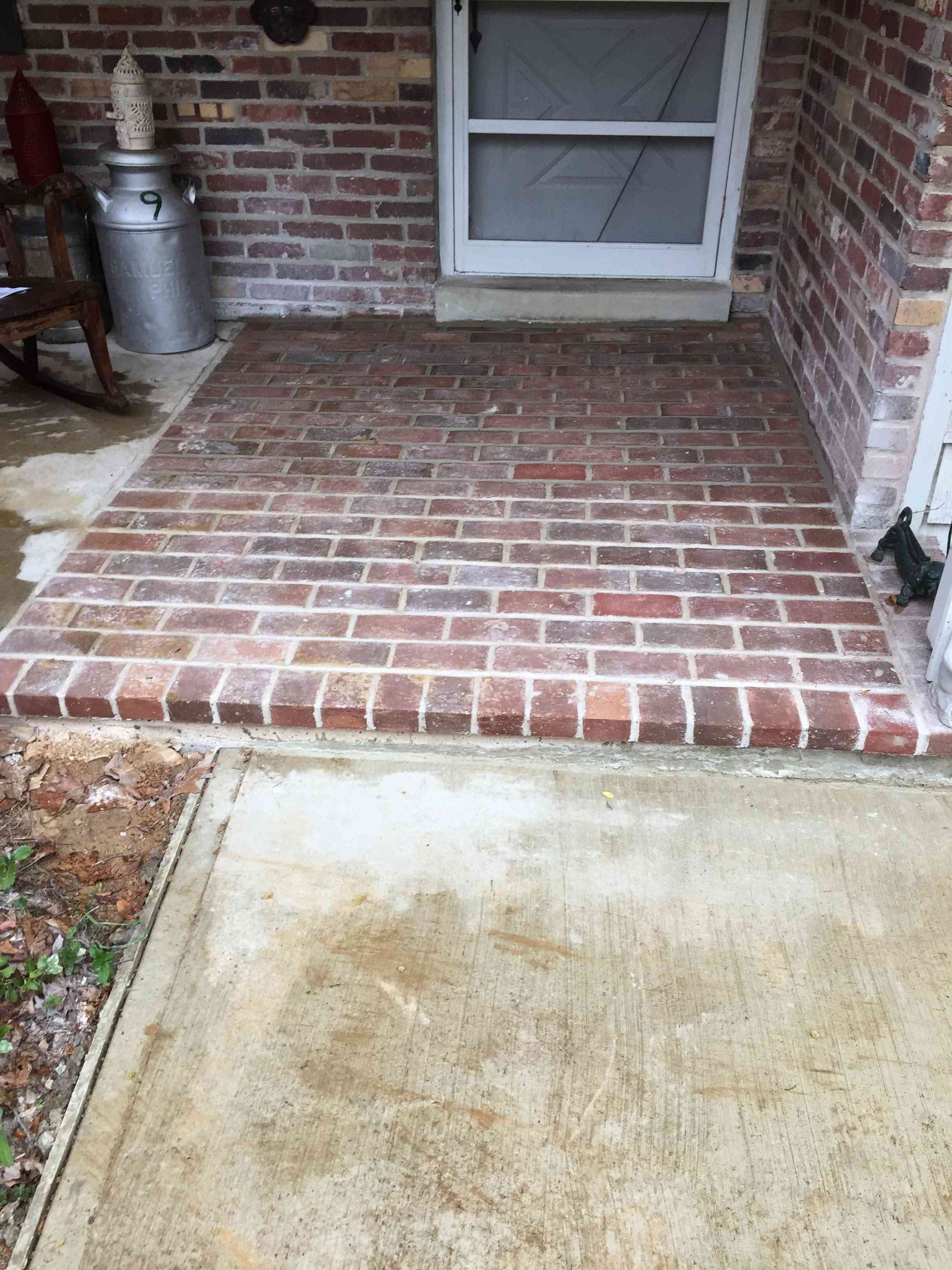 A brick-paved front porch entrance with a metal milk can on the side, leading to a front door.