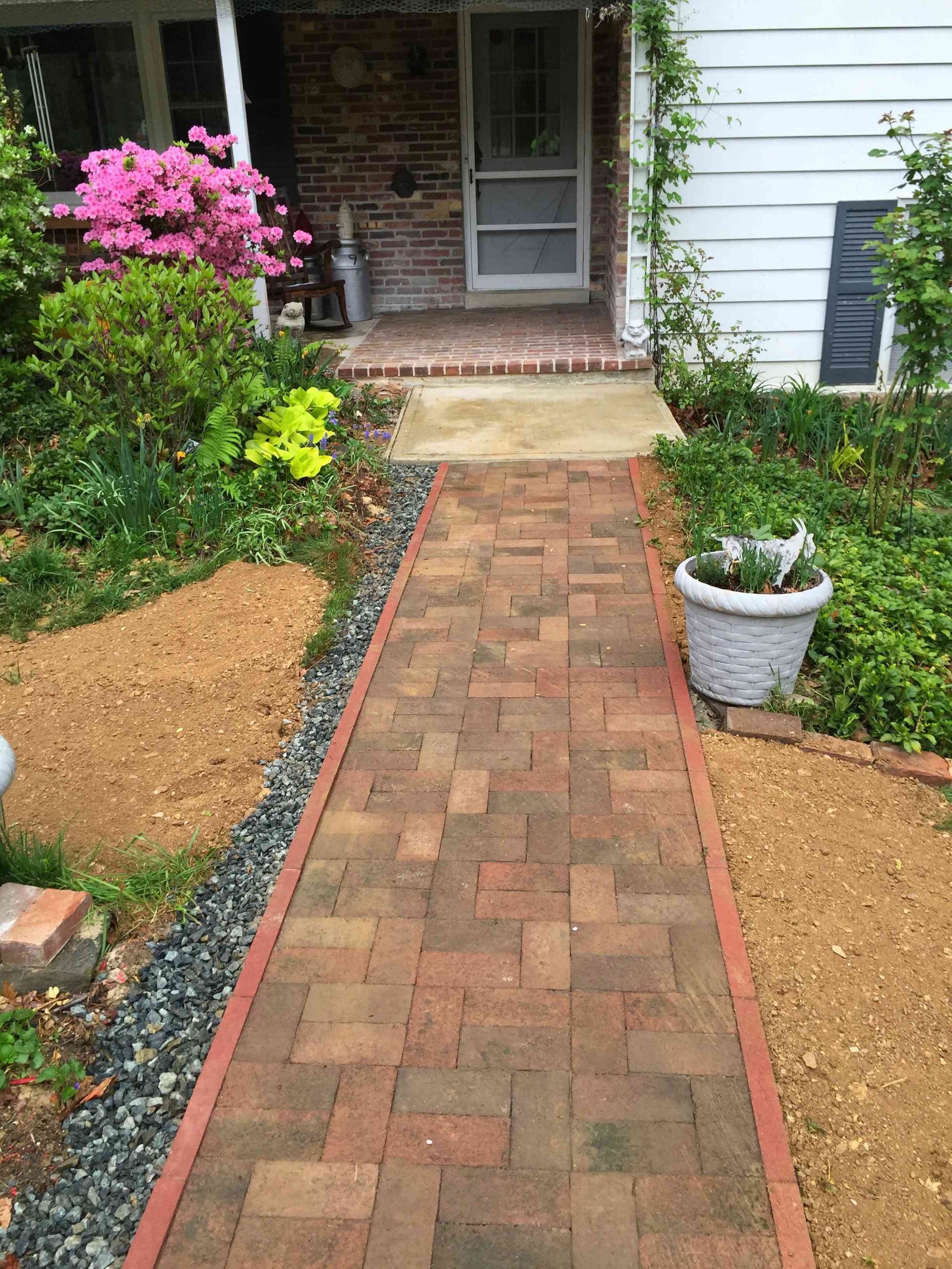A brick walkway leads to the front door of a home, flanked by garden beds with mulch, gravel, and a potted plant.