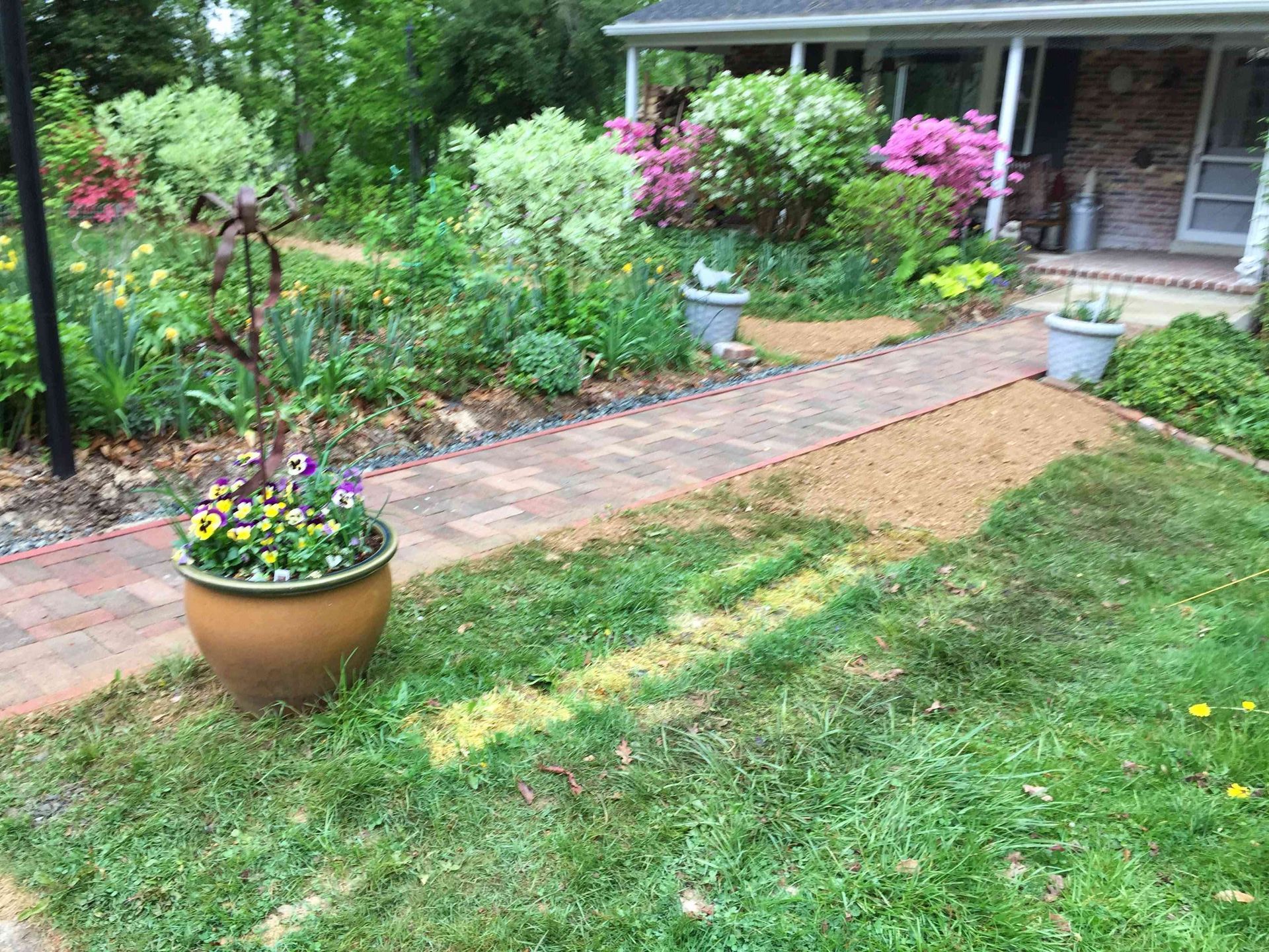 A brick walkway leads to a house entrance, past a flower pot and garden beds filled with blooming pink and green shrubs.