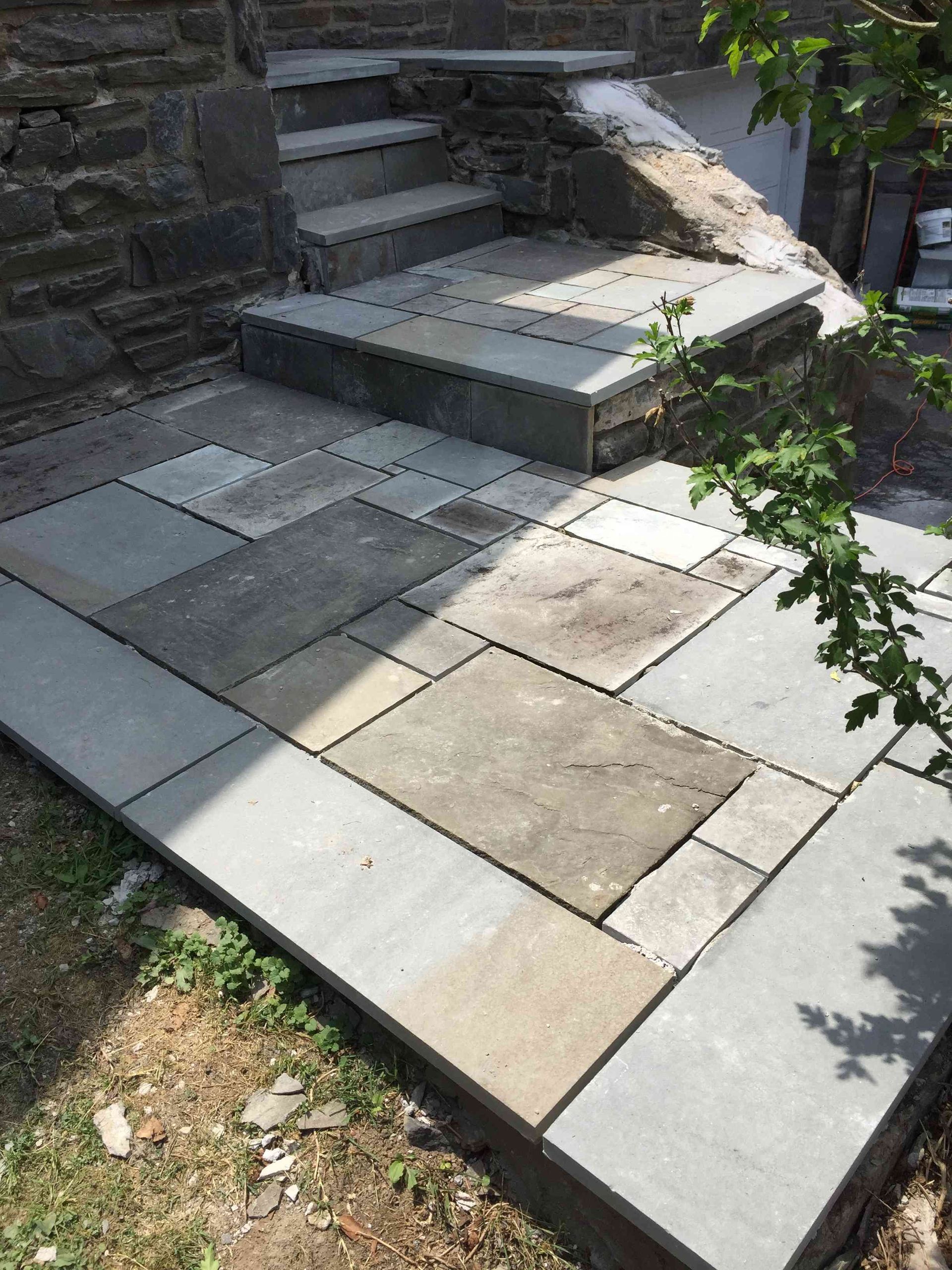 A stone patio with a variety of grey and tan rectangular paving slabs leading to stone steps against a building.