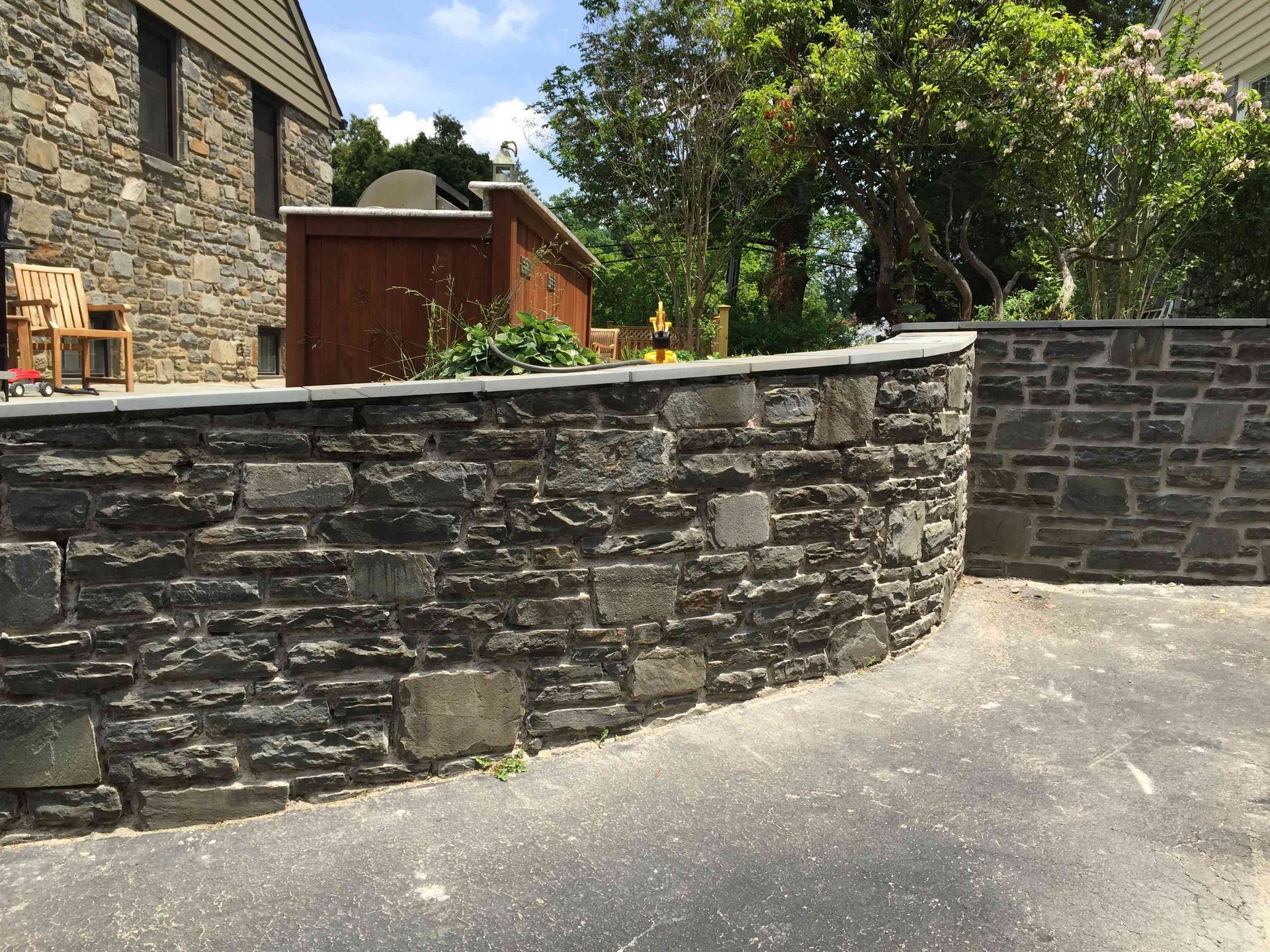 A curved stone retaining wall borders an asphalt driveway next to a stone-clad house.