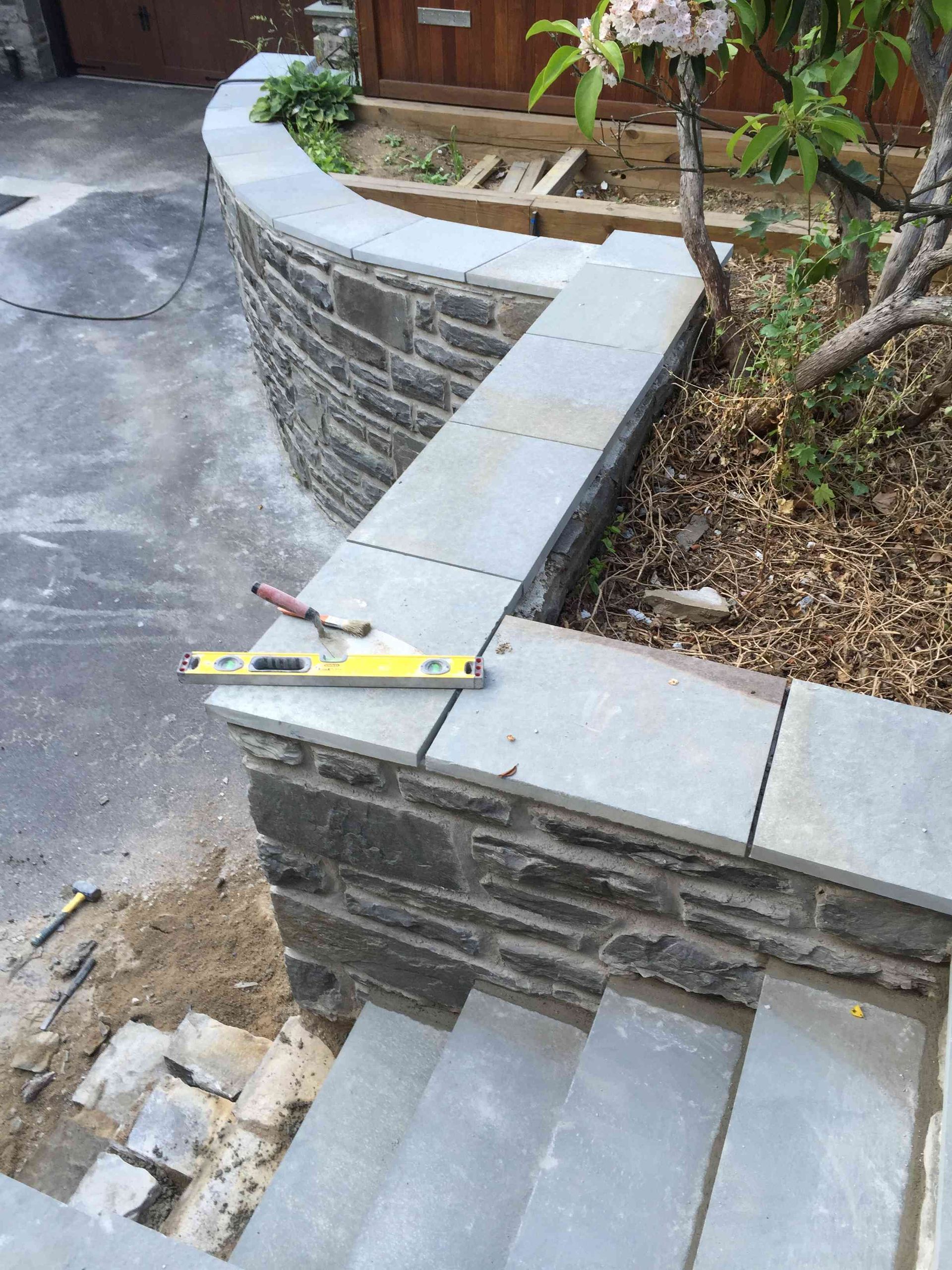 A stone retaining wall with a level and trowel on top, next to a set of stairs under construction in a garden.