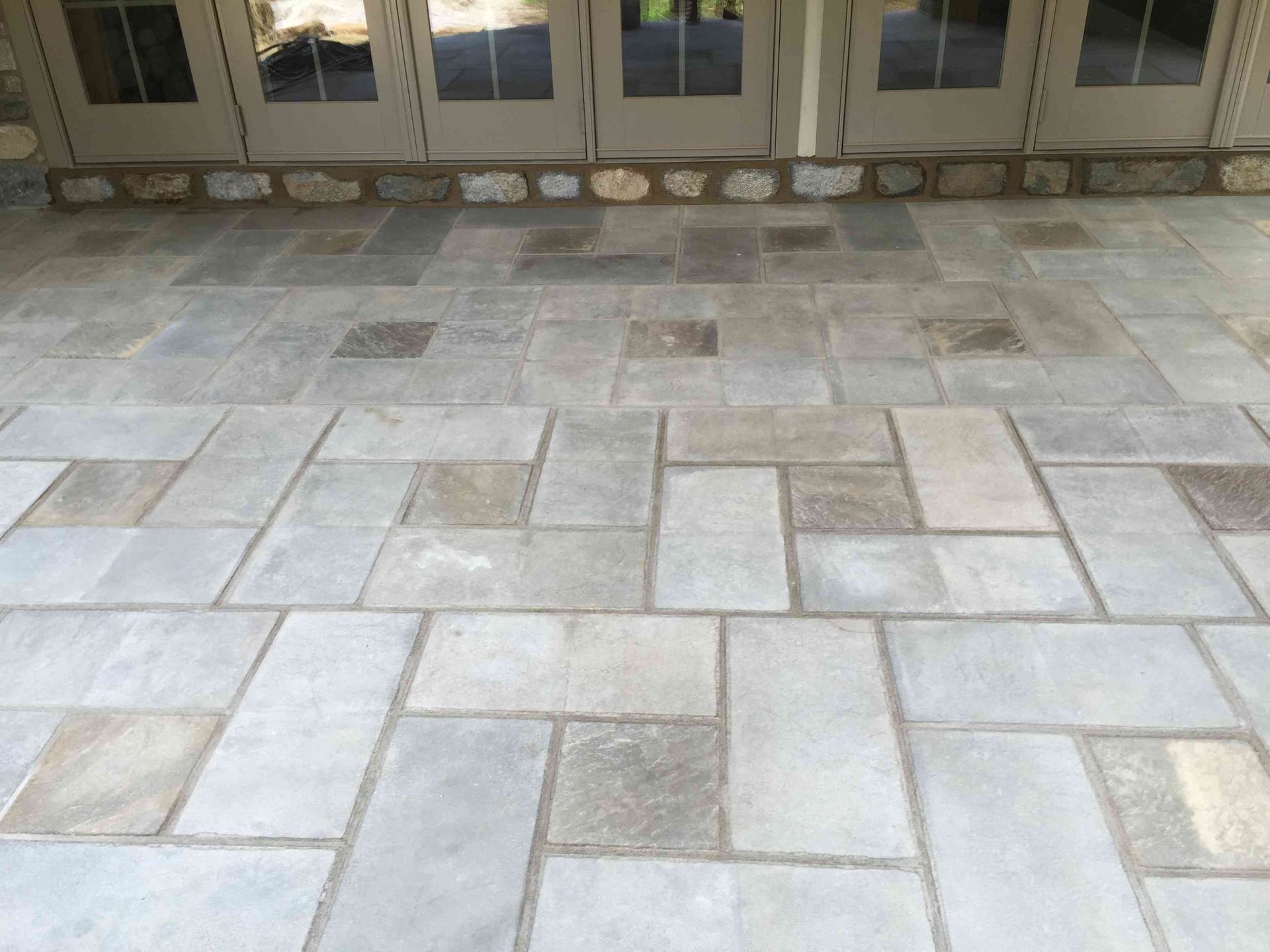 Light gray rectangular stone pavers laid in a repeating, interlocking geometric pattern leading to a set of glass doors.