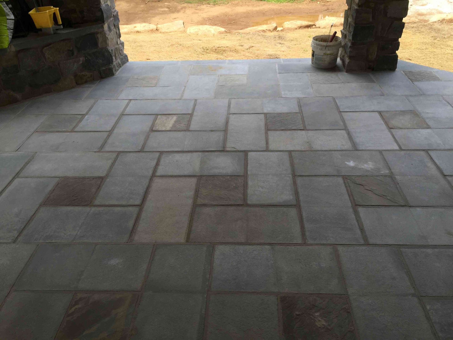 A gray stone patio featuring a repeating rectangular tile pattern, viewed from a porch looking toward a dirt yard.