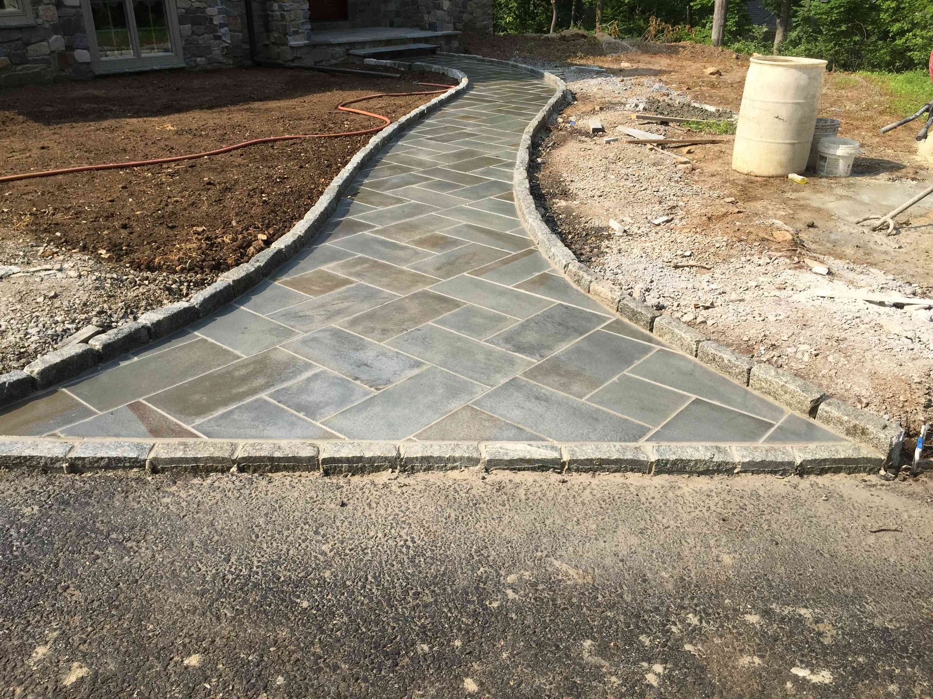 A stone walkway made of gray rectangular pavers in a herringbone pattern with a cobblestone border leads to a house.