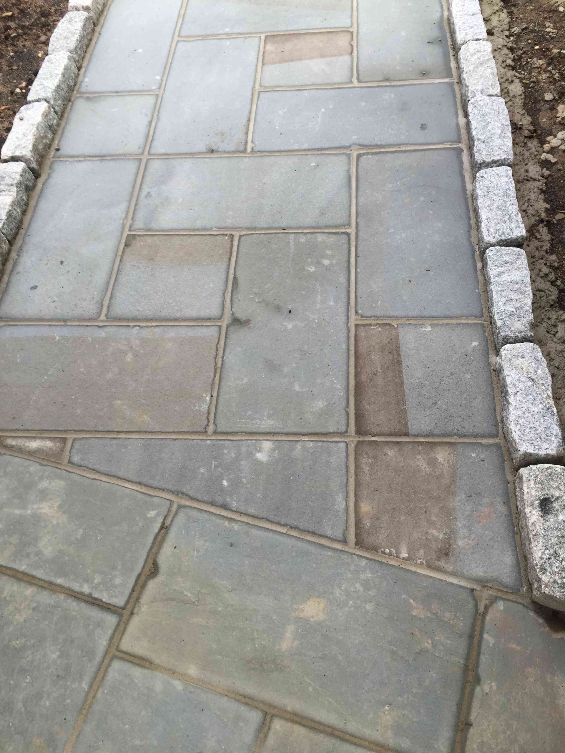 A stone walkway featuring rectangular grey pavers bordered by light-colored, textured granite cobblestones.