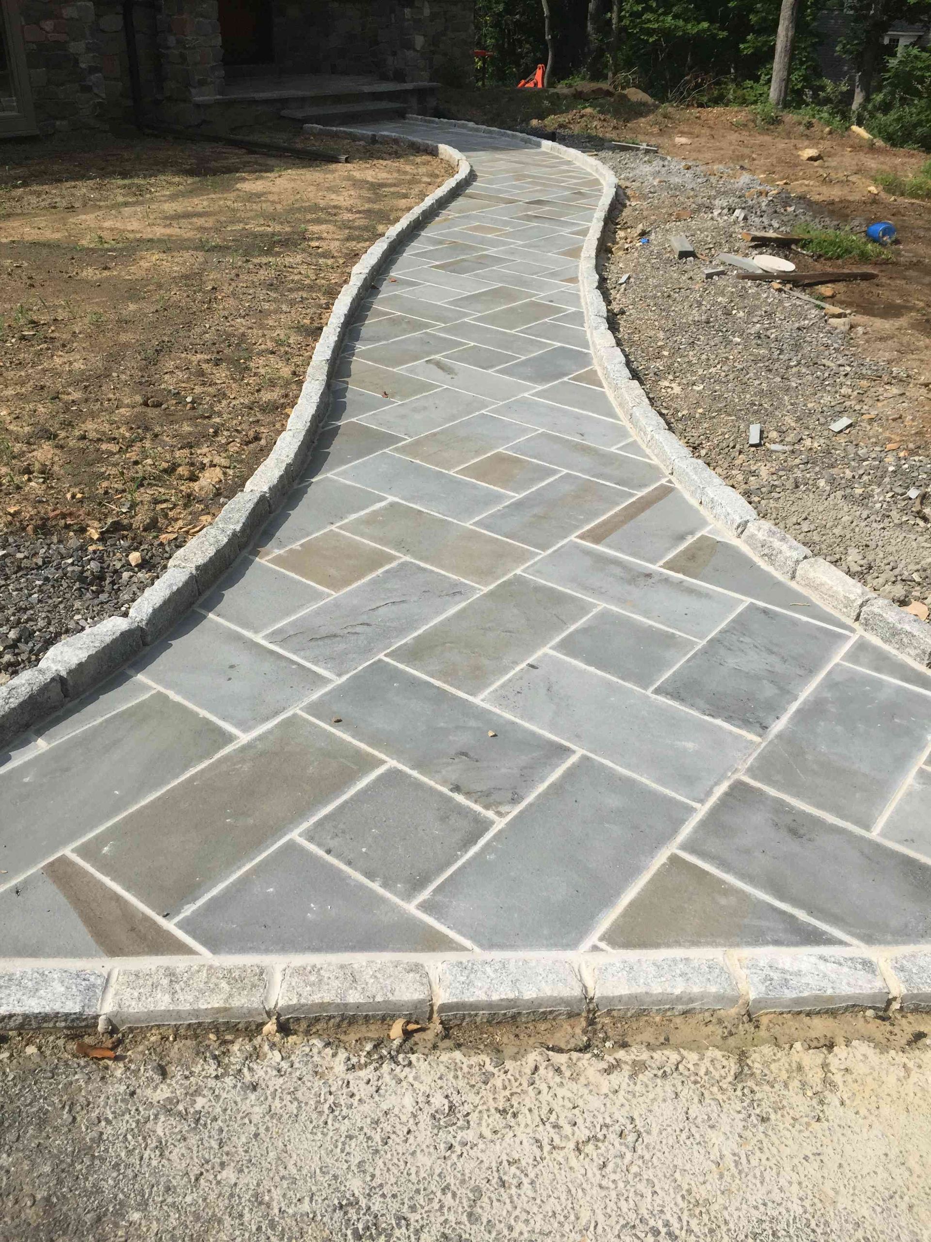 A newly installed walkway made of rectangular gray stone pavers in a herringbone pattern with light stone curbing.