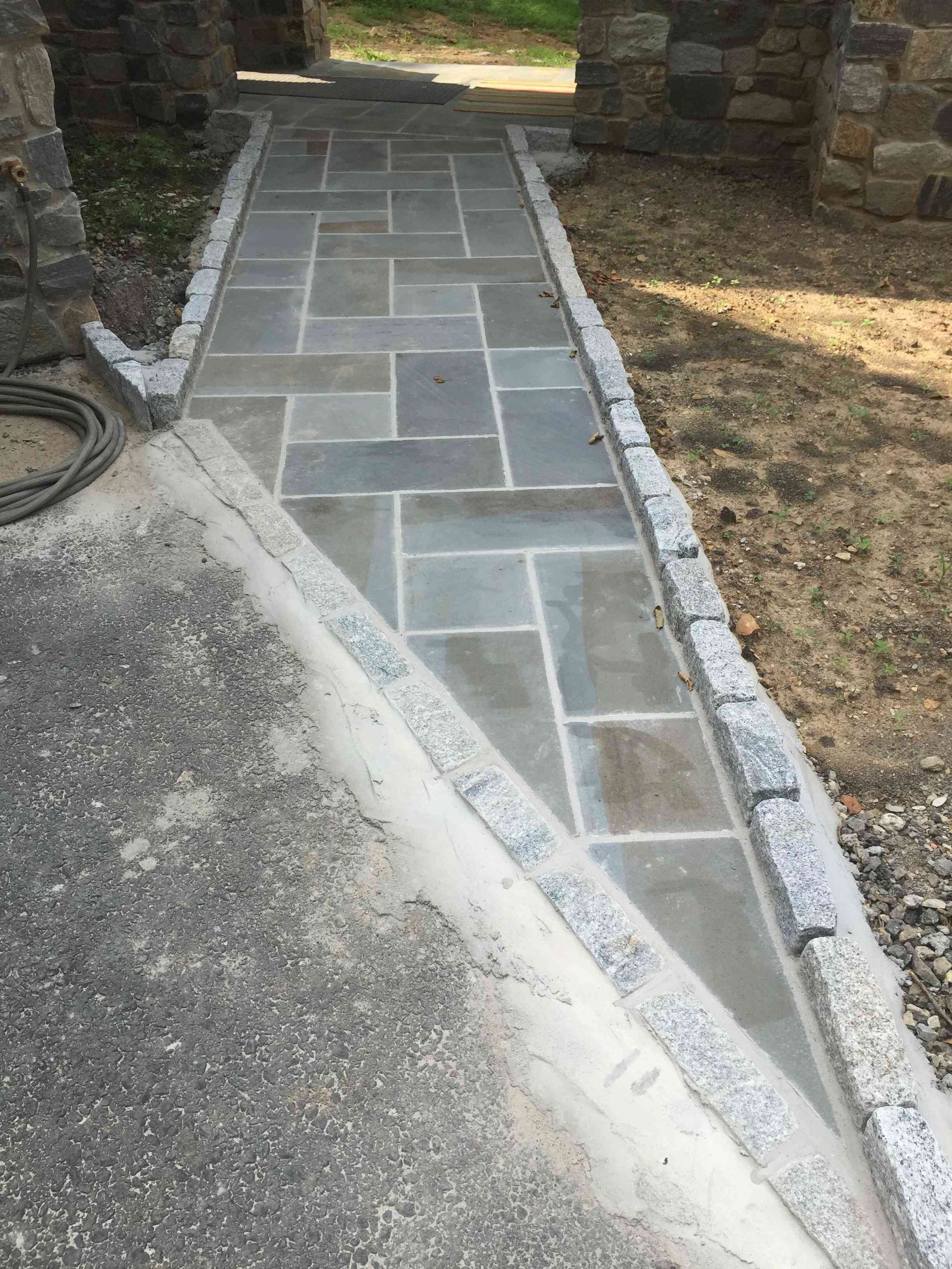 A walkway of rectangular gray pavers bordered by granite blocks, leading from a driveway to a stone building entrance.