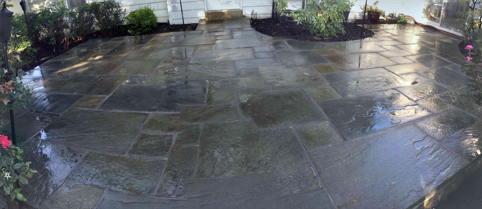 A wide-angle view of a wet stone patio with rectangular pavers in varied gray and tan tones, bordered by garden greenery.