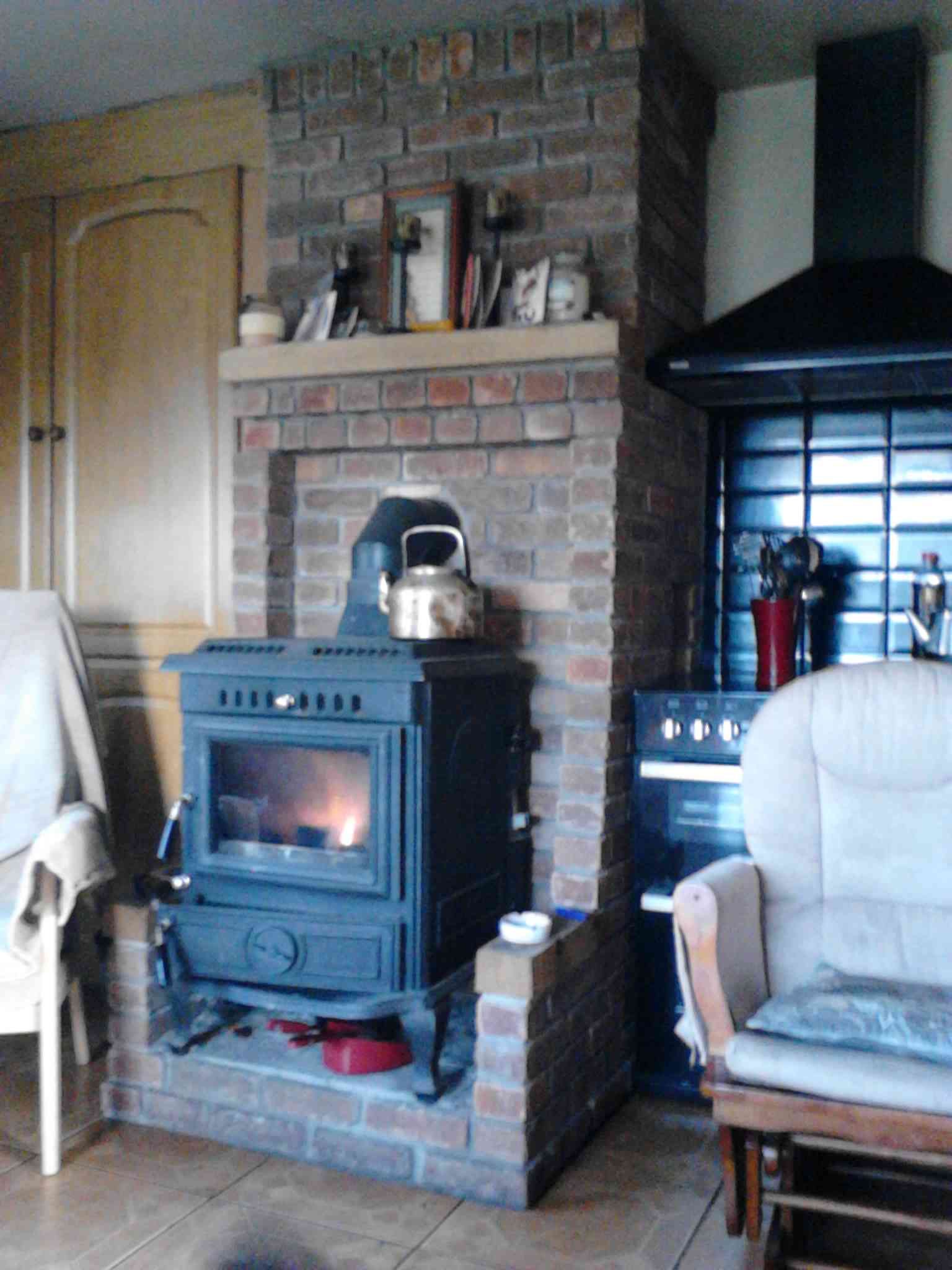 A lit wood-burning stove sits inside a brick fireplace alcove, flanked by two chairs in a room with a black range hood.