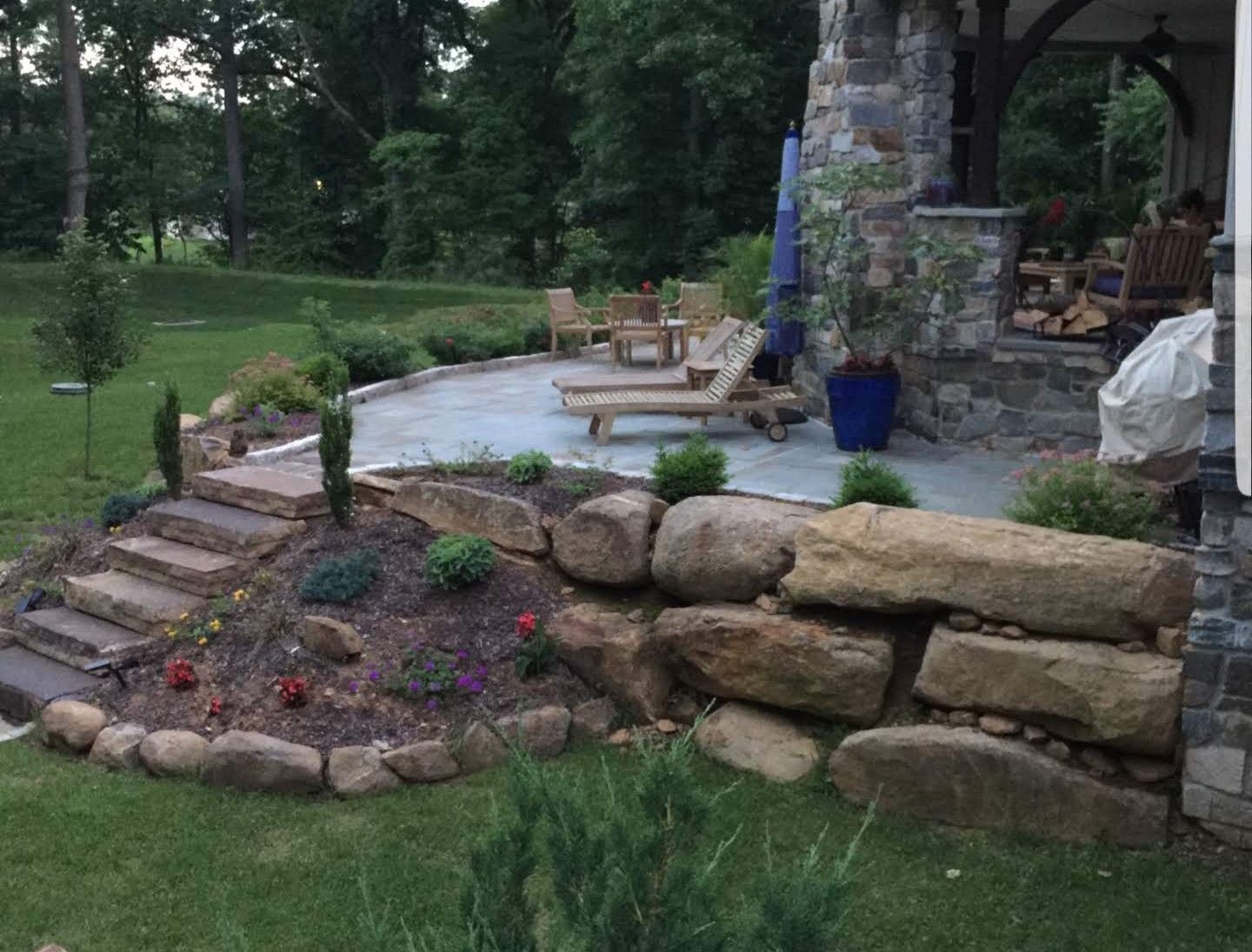 A stone-walled garden bed with steps leads up to a paved patio featuring lounge chairs under a stone-columned porch.