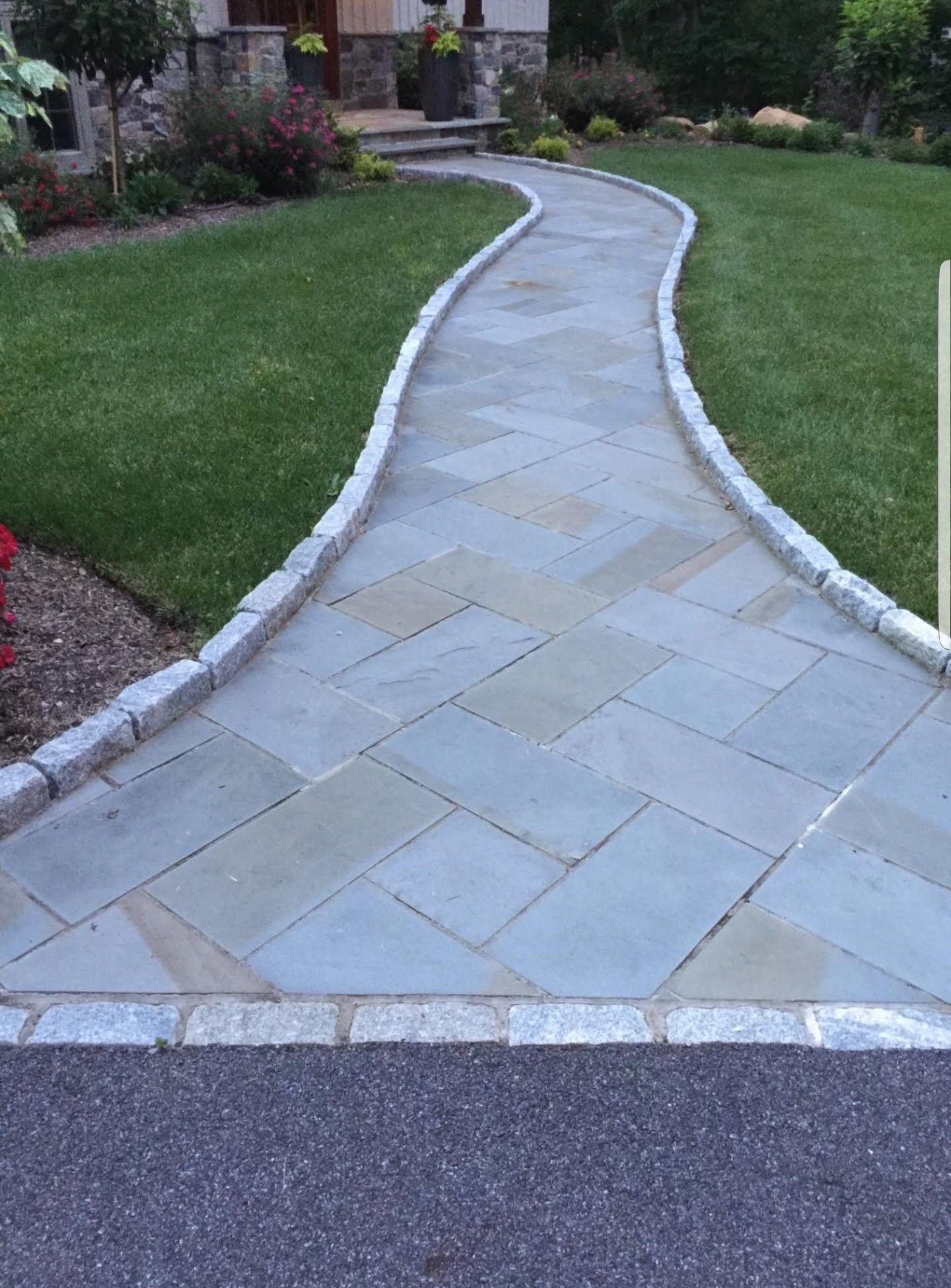 A curved stone paver walkway leading to a house entrance, bordered by grey cobblestones and surrounded by green lawn.