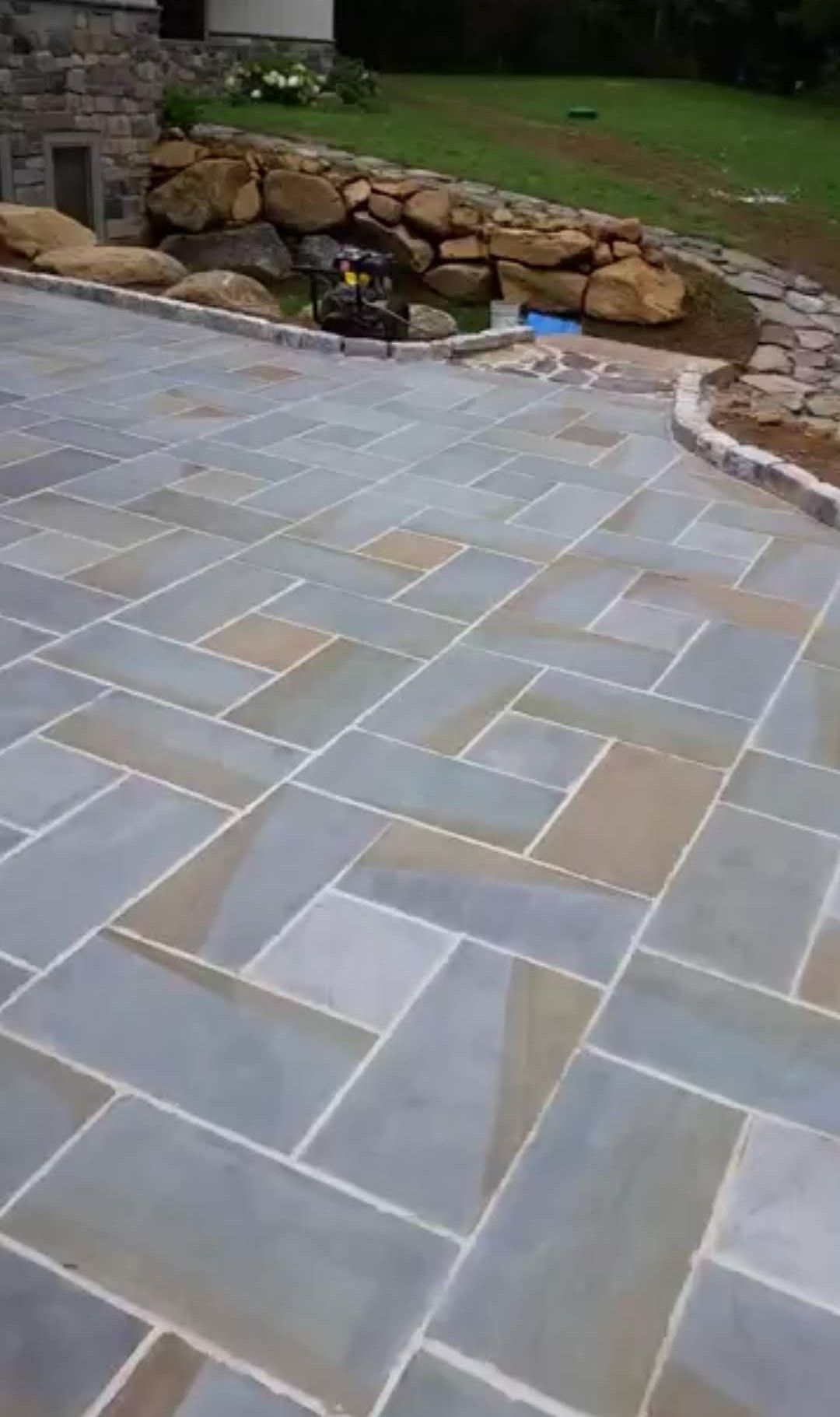 A blue and tan stone patio with a pinwheel pattern, bordered by a low rock wall and grass.