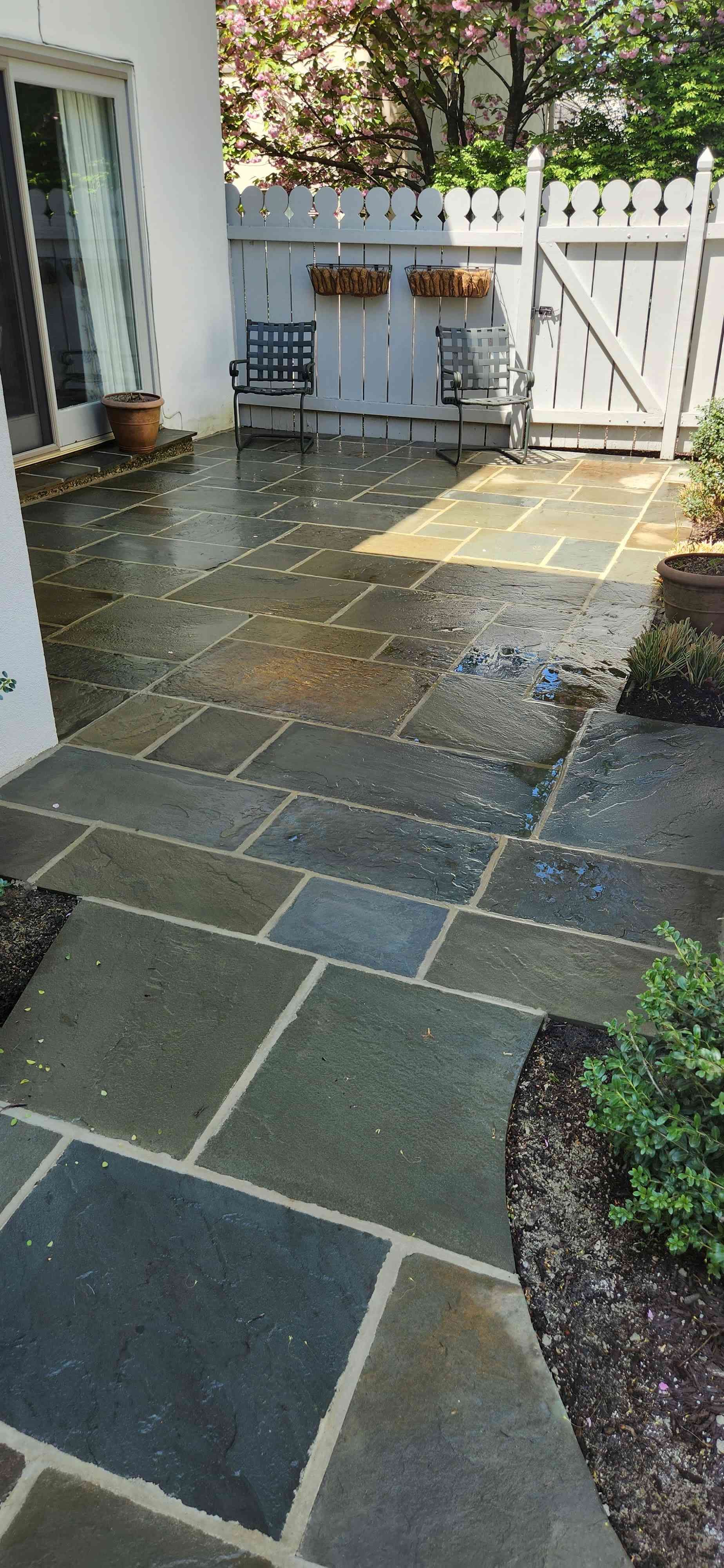 A patio featuring wet, multi-colored stone pavers in a random pattern, with a white fence, chairs, and plants nearby.