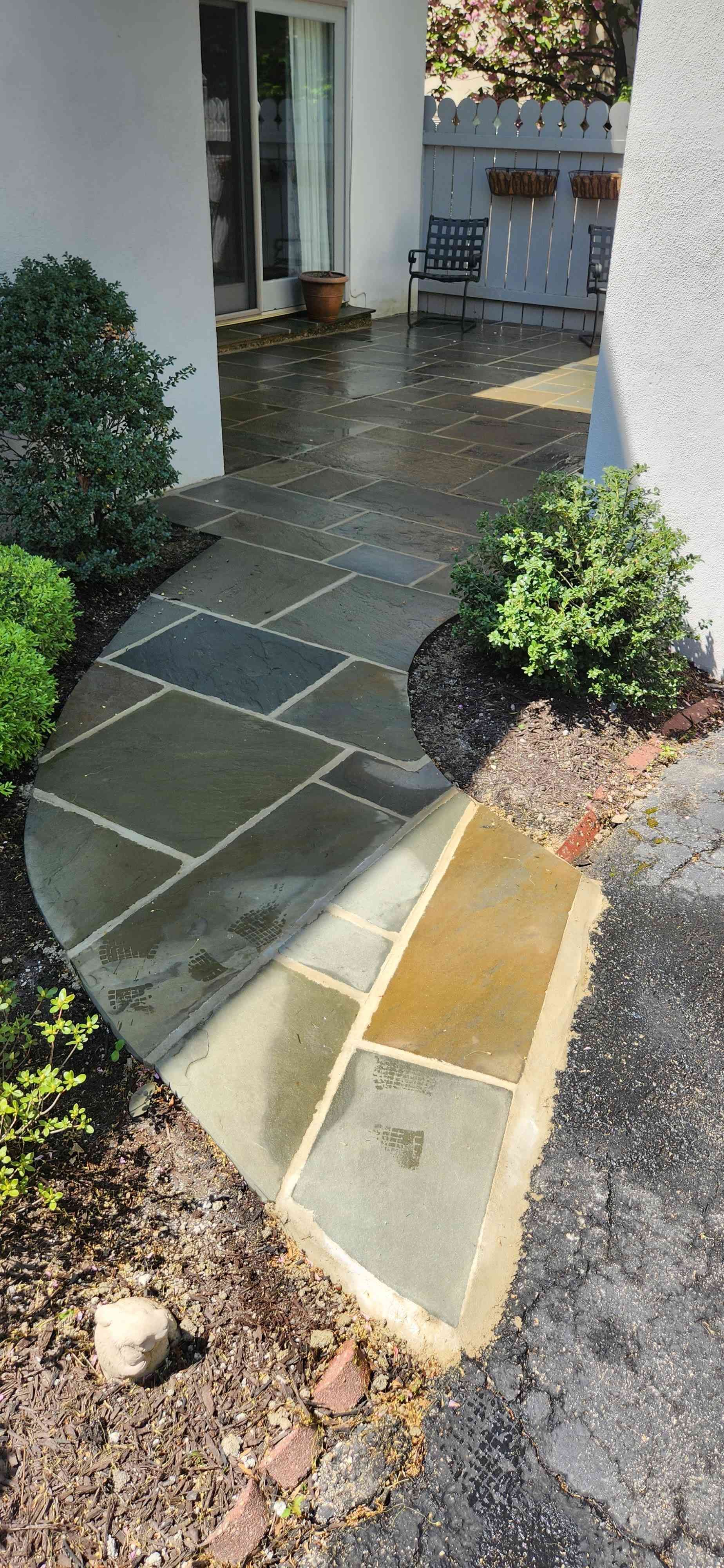 A stone pathway leads from a driveway to a patio with sliding glass doors, framed by green shrubs and mulch beds.