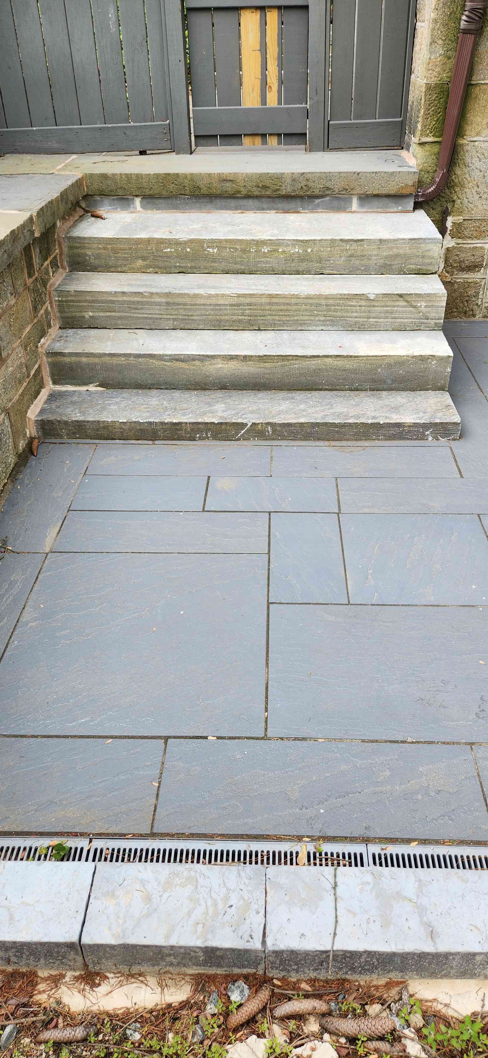 A set of three grey stone steps leading up to a wooden gate, set against a patio area with a dark drainage channel.