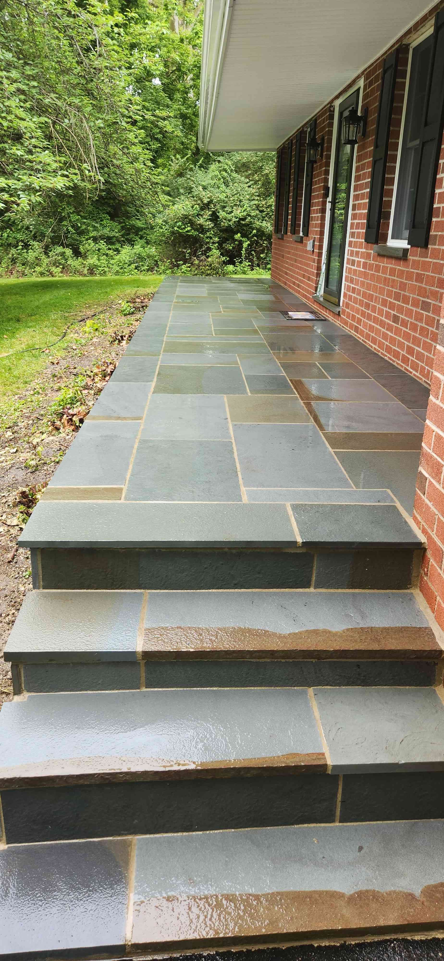 A stone-tiled porch and set of stairs leading to a brick house entrance, with green trees and landscaping in the background.
