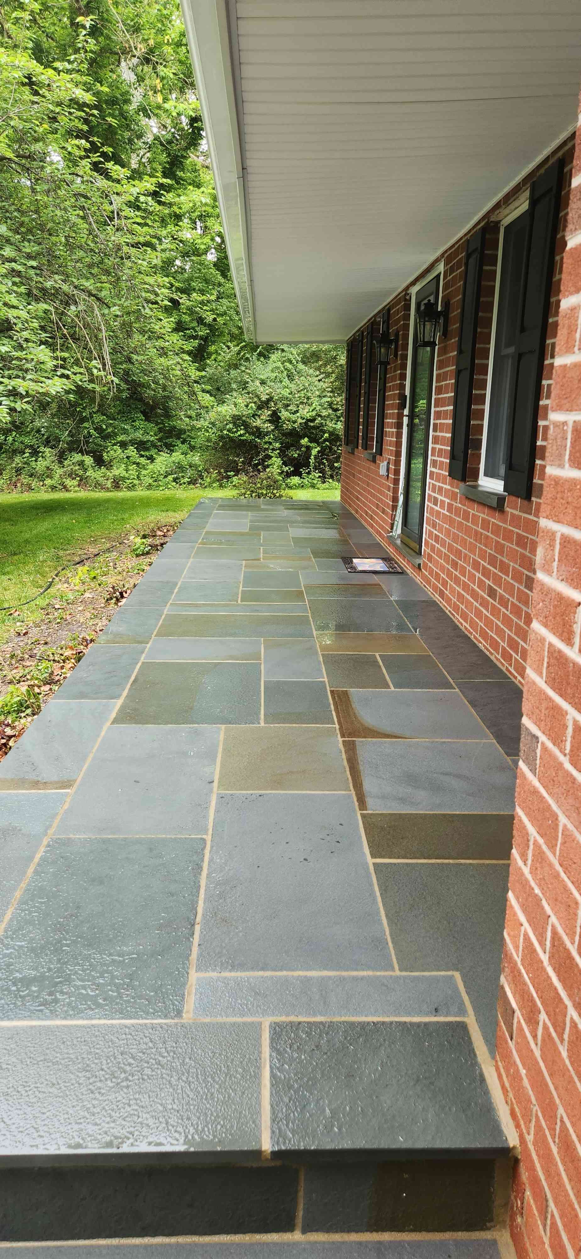 A blue-gray slate walkway leads along a red brick house with dark shutters, flanked by a lawn and trees.