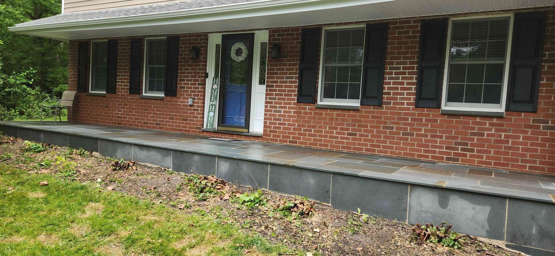 A red brick house with a blue front door, black shutters, and a long, grey stone front porch with landscaping.