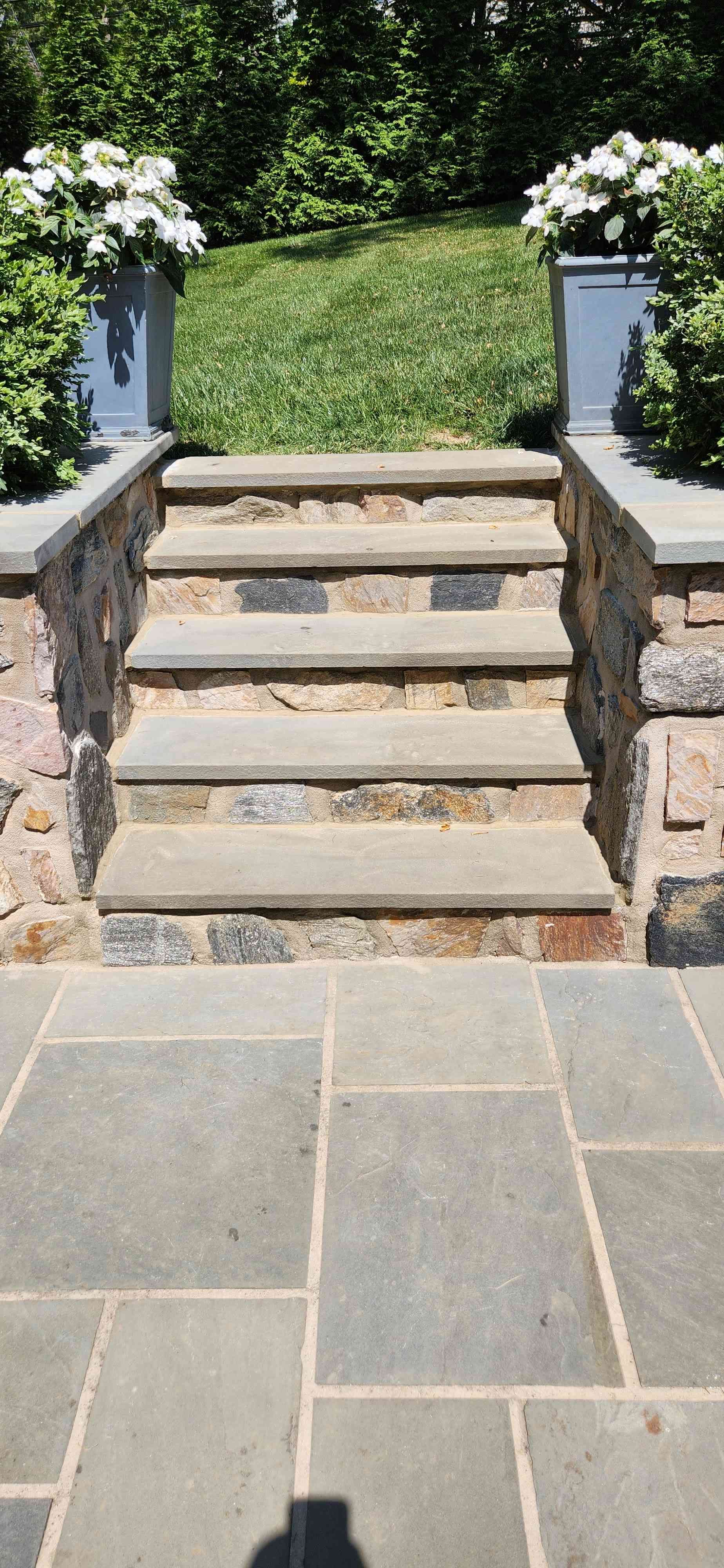 Stone steps with flagstone treads leading to a grassy hill between two planters with white flowers.