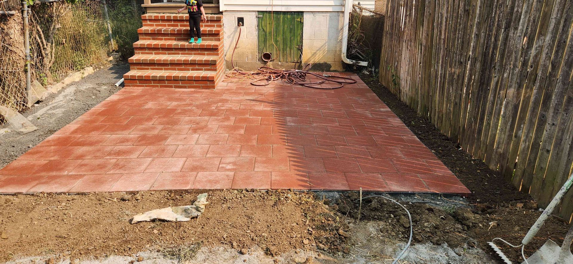 A rectangular red brick patio sits in a backyard next to wooden stairs and a wooden fence, with unfinished landscaping.