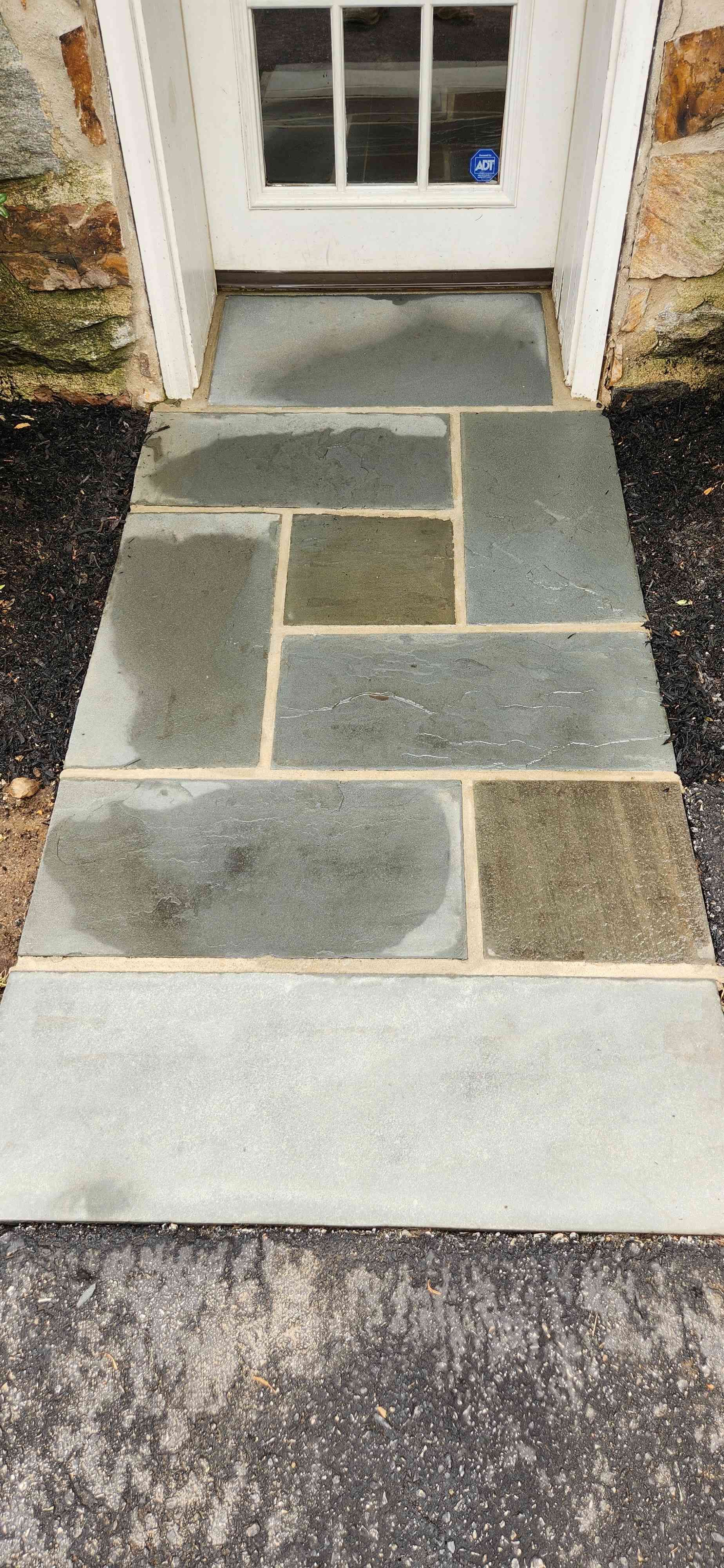 A stone walkway with a rectangular, mosaic-like pattern leads to a white door framed by stone walls.