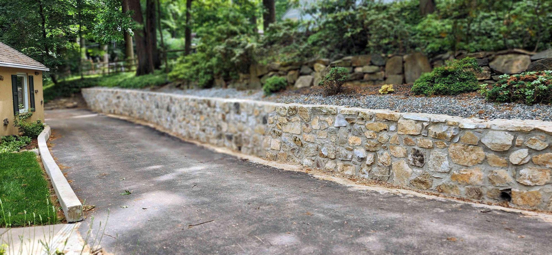 A long, tan stone retaining wall runs alongside a paved driveway next to a grassy slope and a portion of a house.