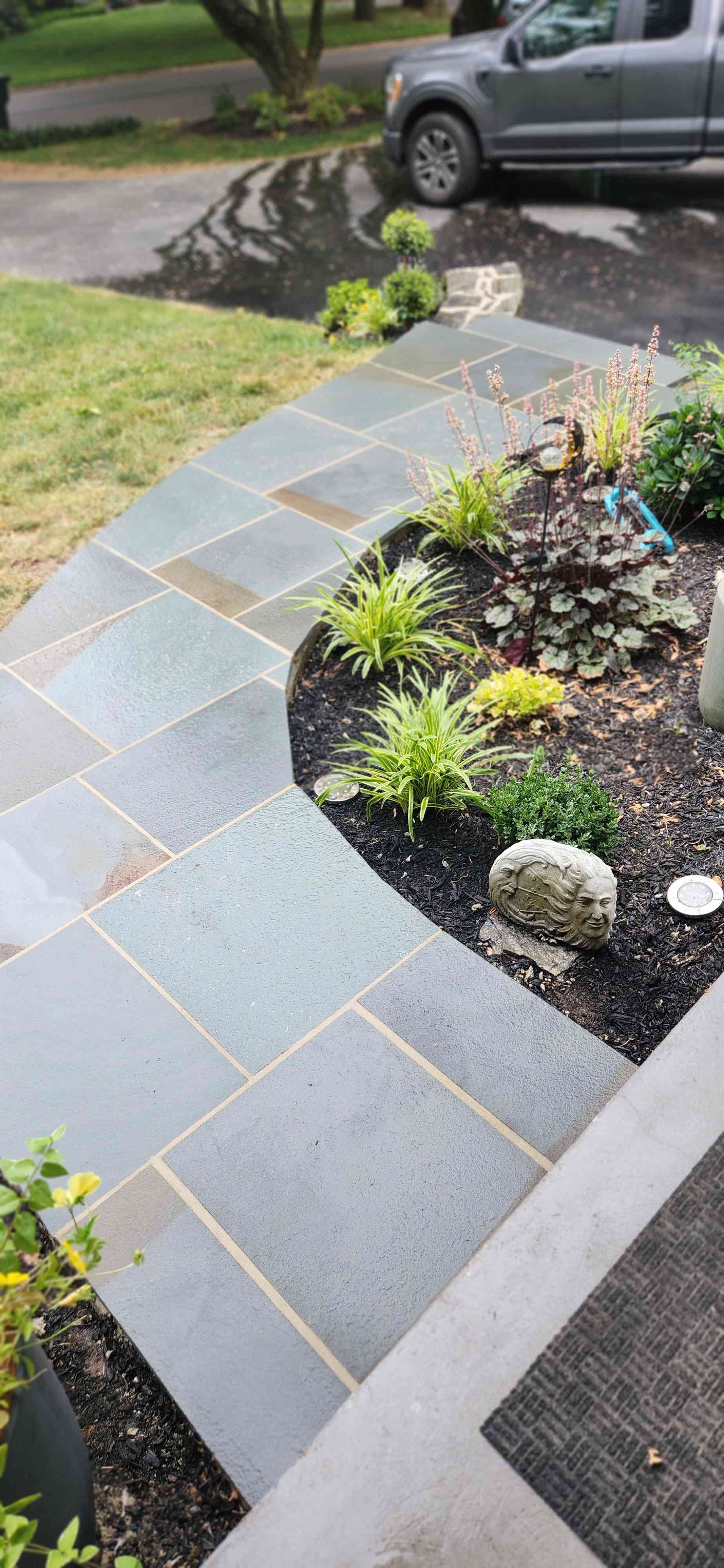 A curved path of square, gray stone pavers leading to a home entrance, adjacent to a mulched garden bed with small plants.