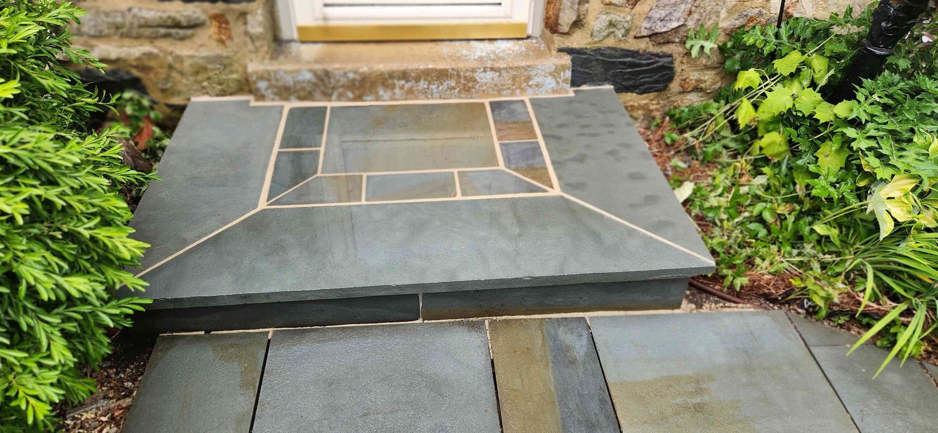 A stone front porch entrance with a rectangular inlay pattern, surrounded by greenery and a stone foundation.