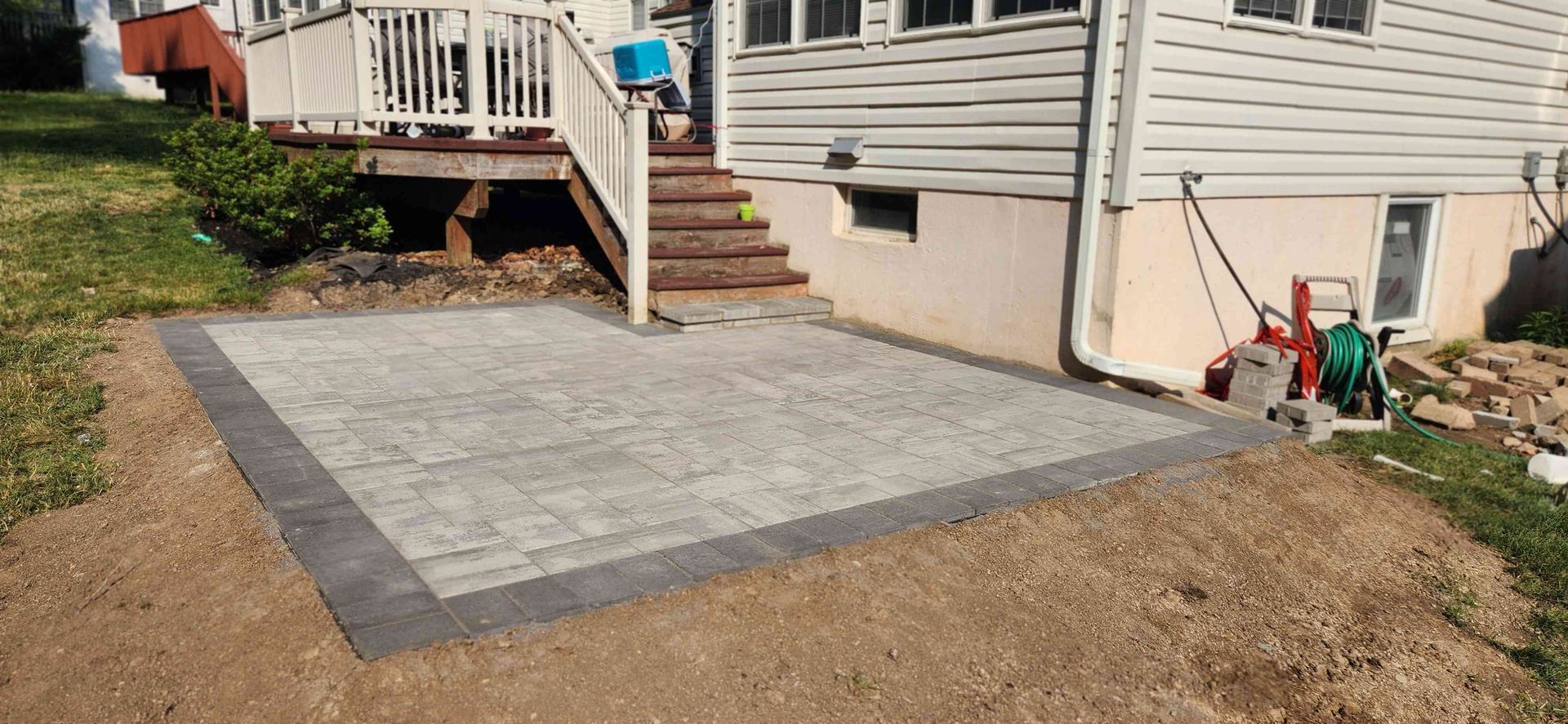 A newly installed rectangular stone patio with a dark border sits next to a house with stairs leading to a wood deck.
